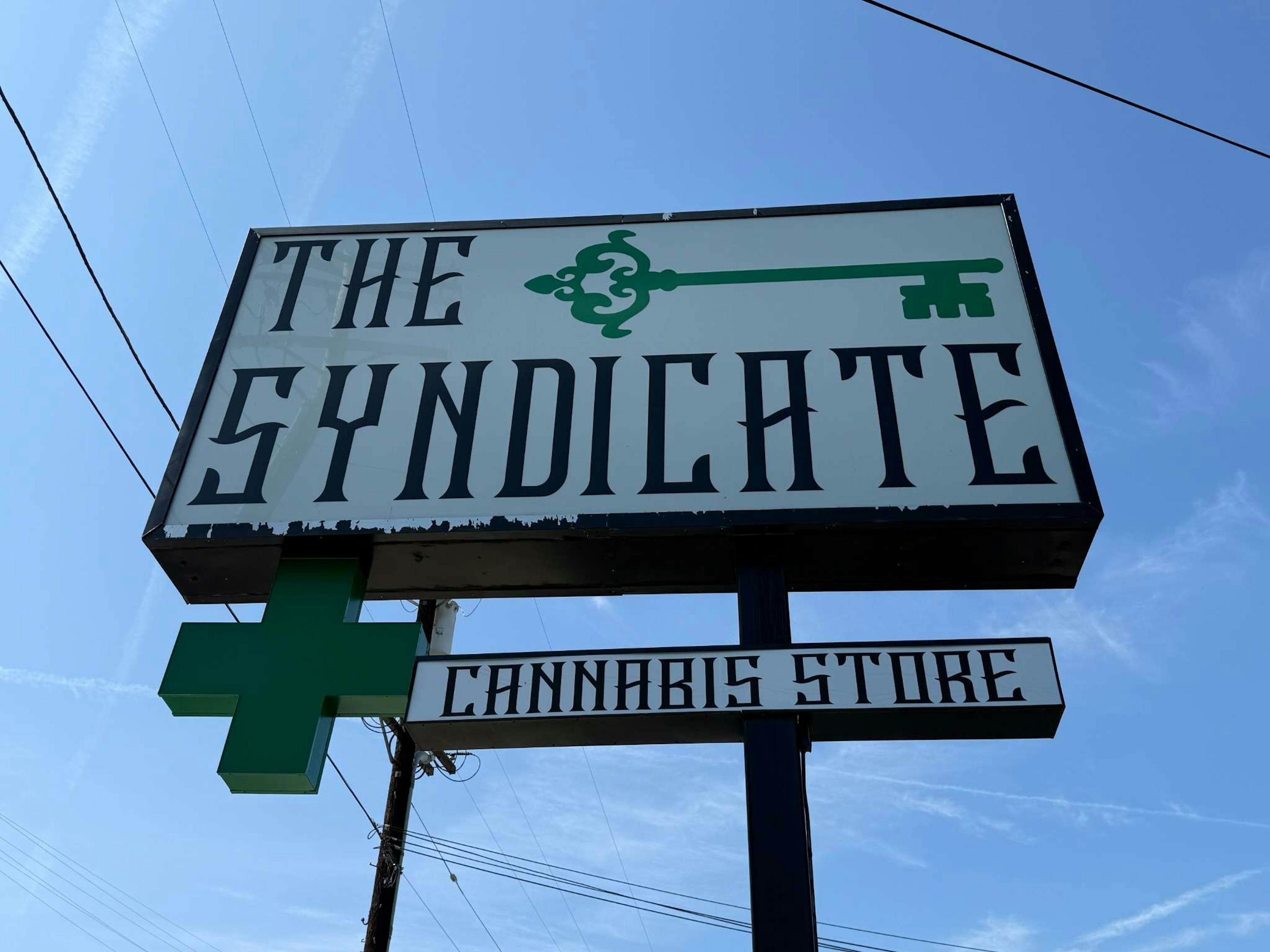 Tall shot of The Syndicate’s road sign prominently displaying the green key logo and “Cannabis Store” label.