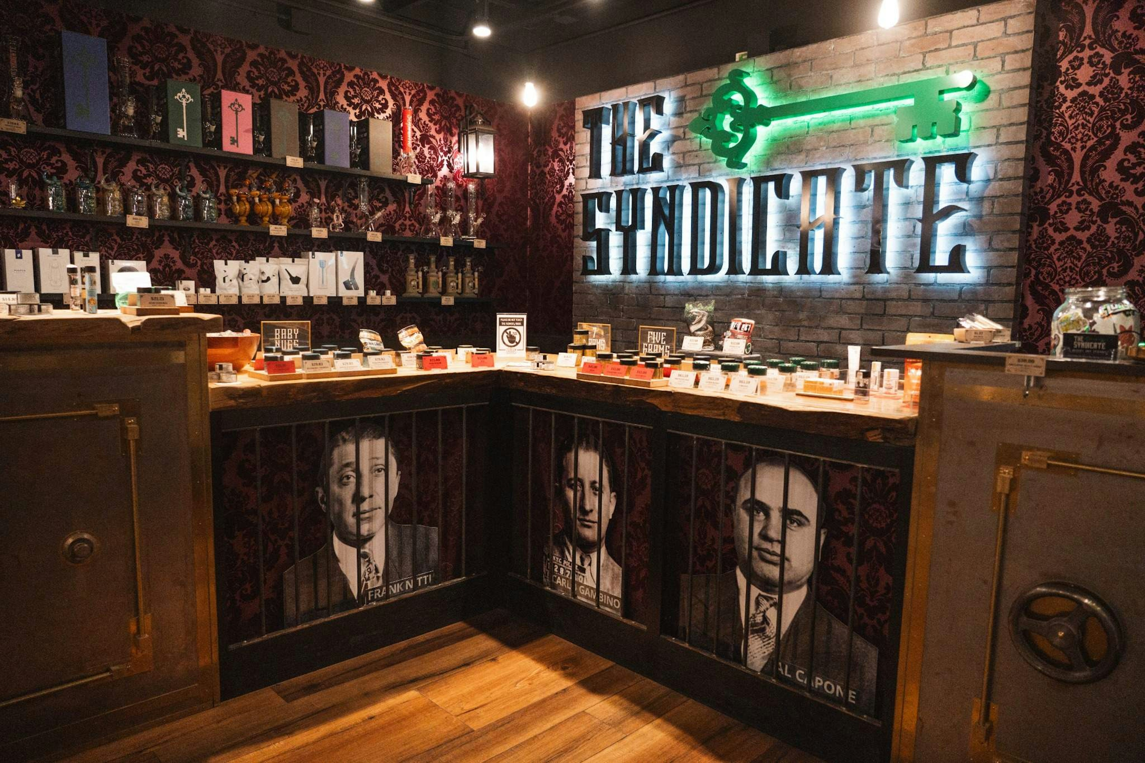 Interior of The Syndicate dispensary featuring a vintage-style counter with portraits of historic mob figures Frank Nitti, Carlo Gambino, and Al Capone, beneath a glowing neon sign reading “The Syndicate” against a brick wall backdrop. Various cannabis products and accessories are displayed on the counter and shelves.