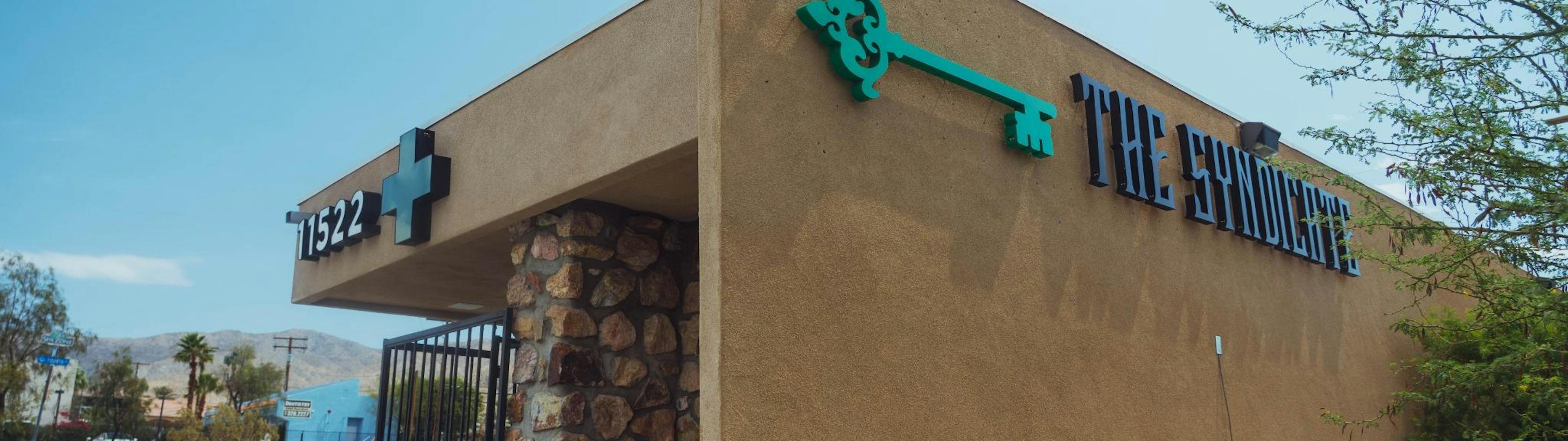 Corner angle of The Syndicate dispensary exterior with the address number 11522 and a medical cannabis cross symbol, green key logo, and decorative stone column under a clear sky, with desert landscape in the background.