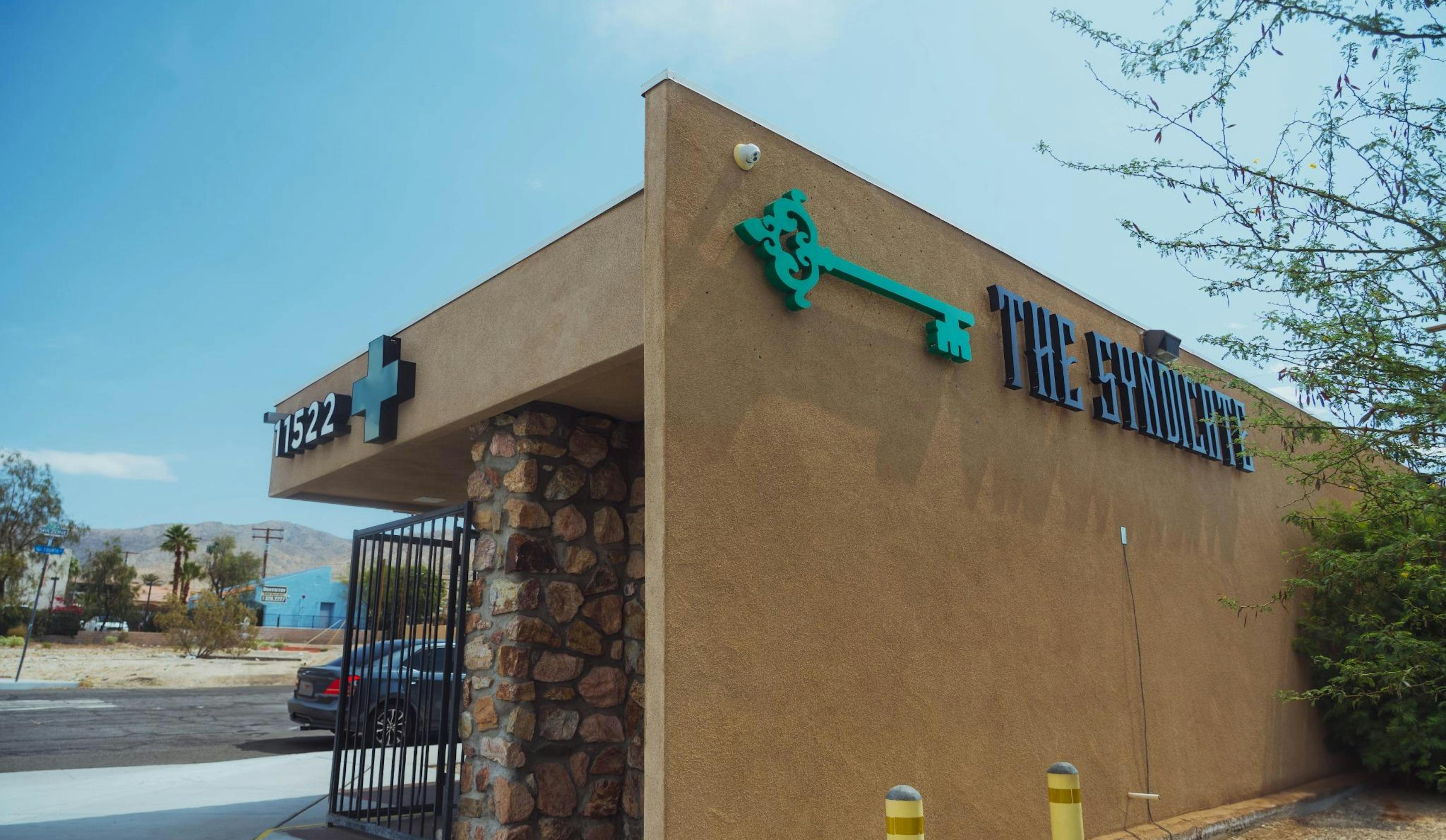 Corner angle of The Syndicate dispensary exterior with the address number 11522 and a medical cannabis cross symbol, green key logo, and decorative stone column under a clear sky, with desert landscape in the background.