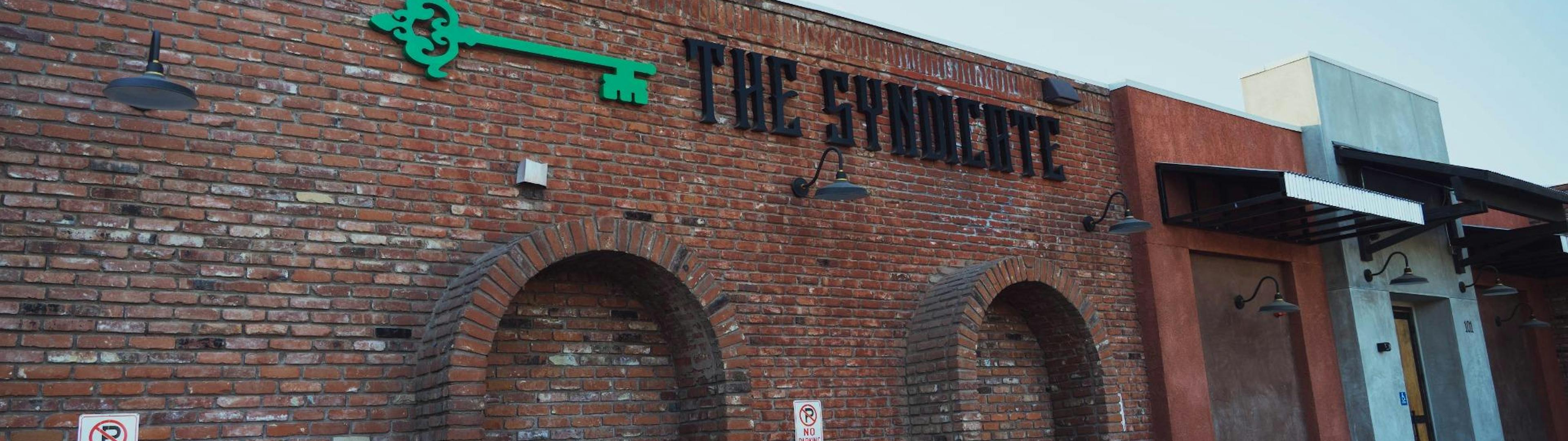Exterior shot of The Syndicate cannabis store in Riverside with red brick walls, signage, and designated parking signage.