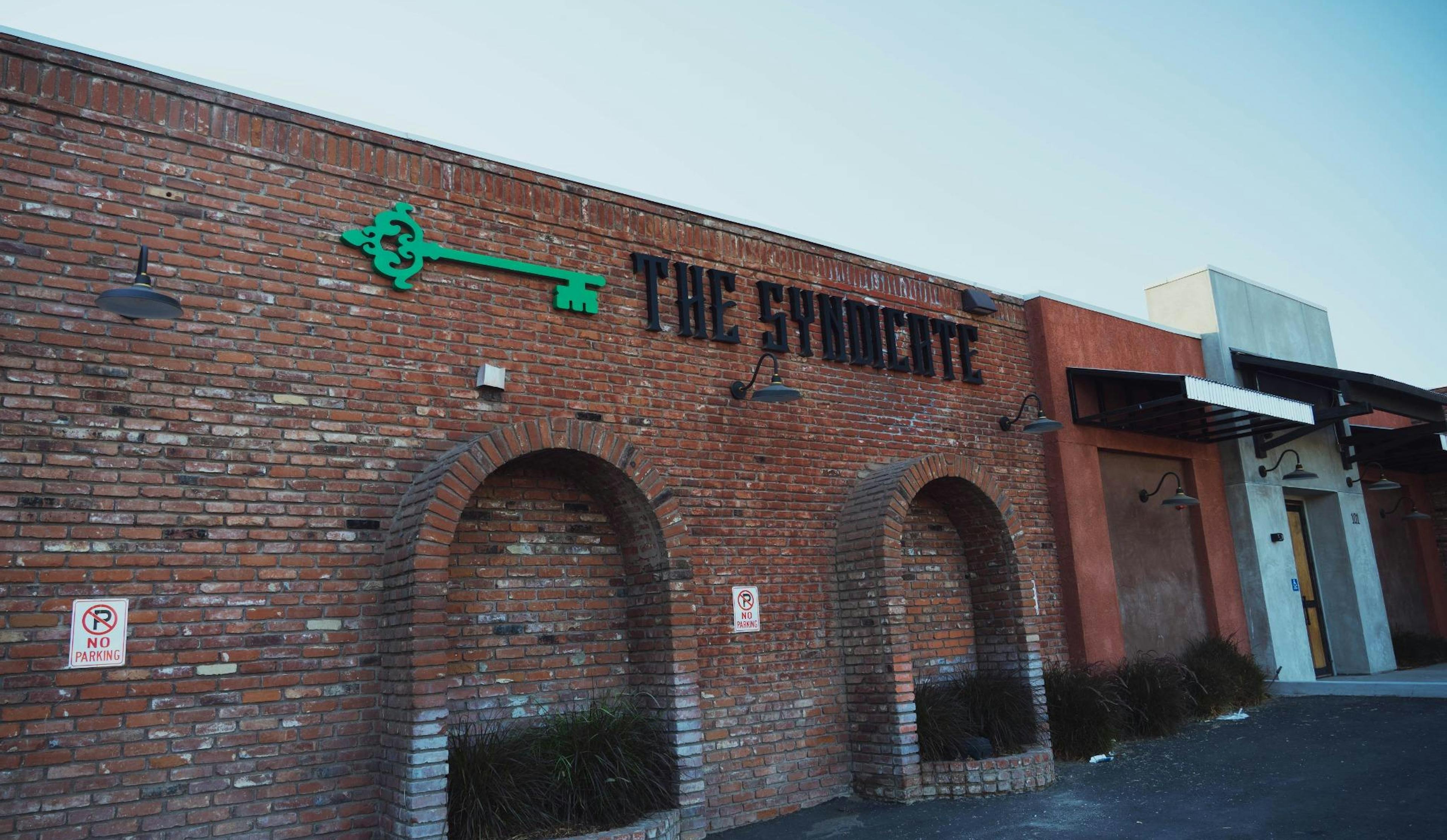Exterior shot of The Syndicate cannabis store in Riverside with red brick walls, signage, and designated parking signage.