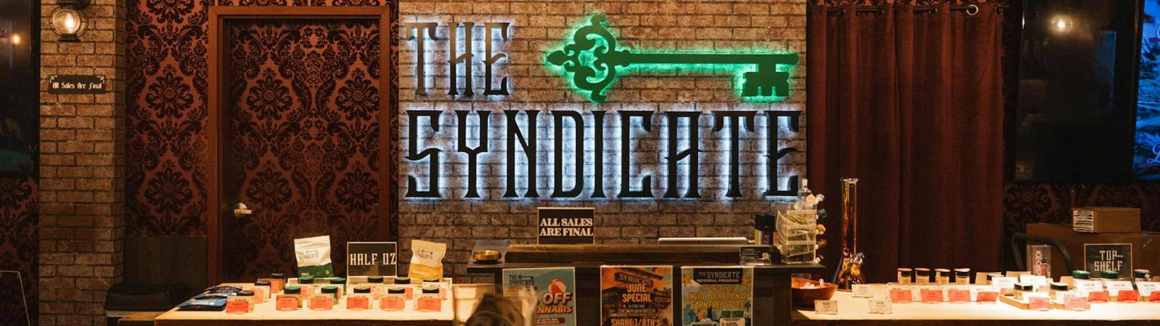 Front counter view at The Syndicate Lake Elsinore dispensary with glowing signage and promotional posters for cannabis products