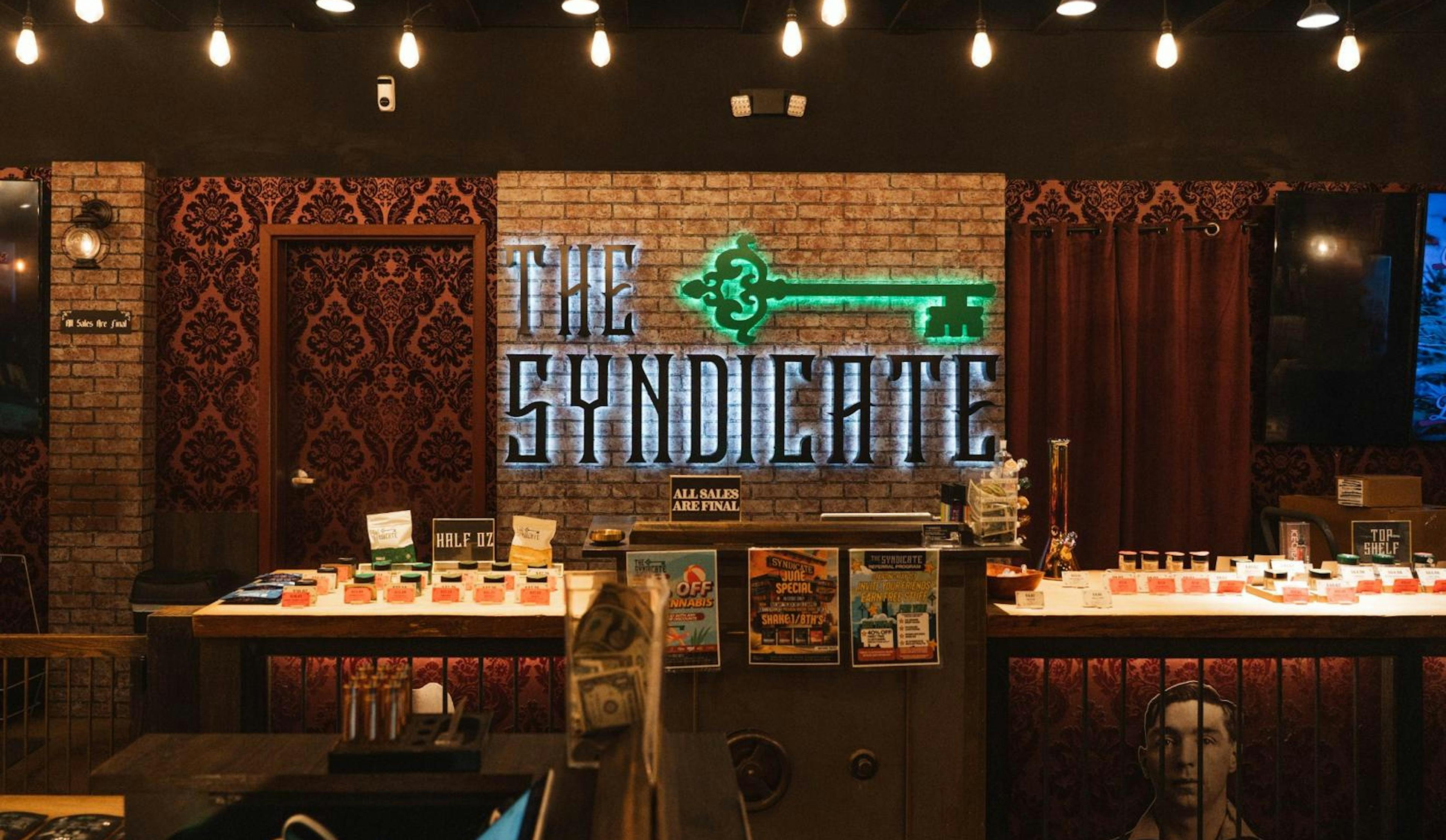 Front counter view at The Syndicate Lake Elsinore dispensary with glowing signage and promotional posters for cannabis products