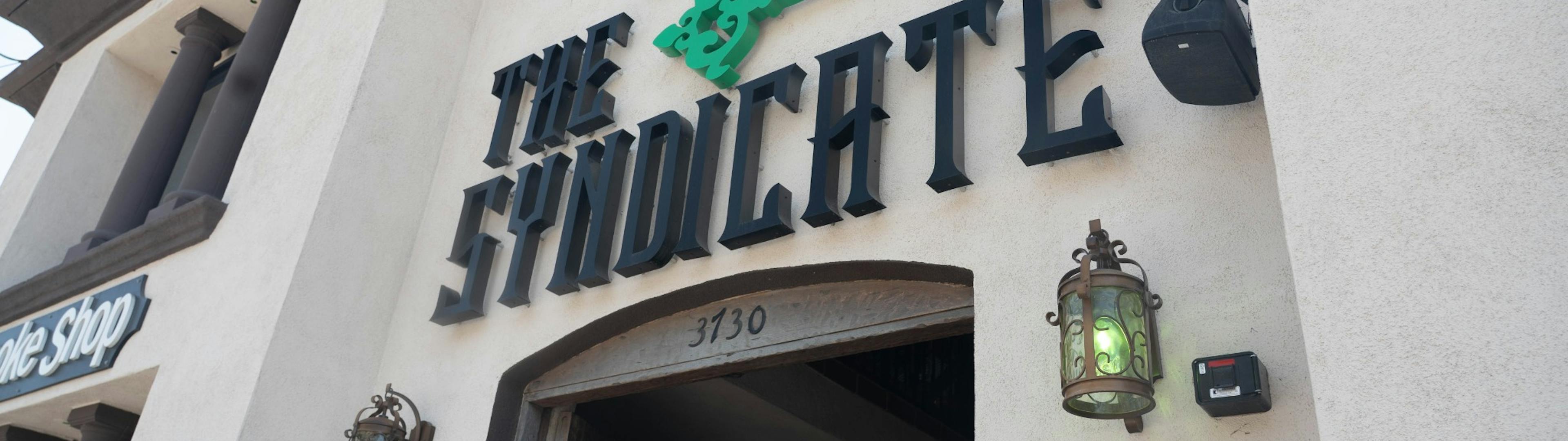 Angled upward view of The Syndicate Long Beach storefront showing the logo with green key and ornate lantern light fixtures above the door