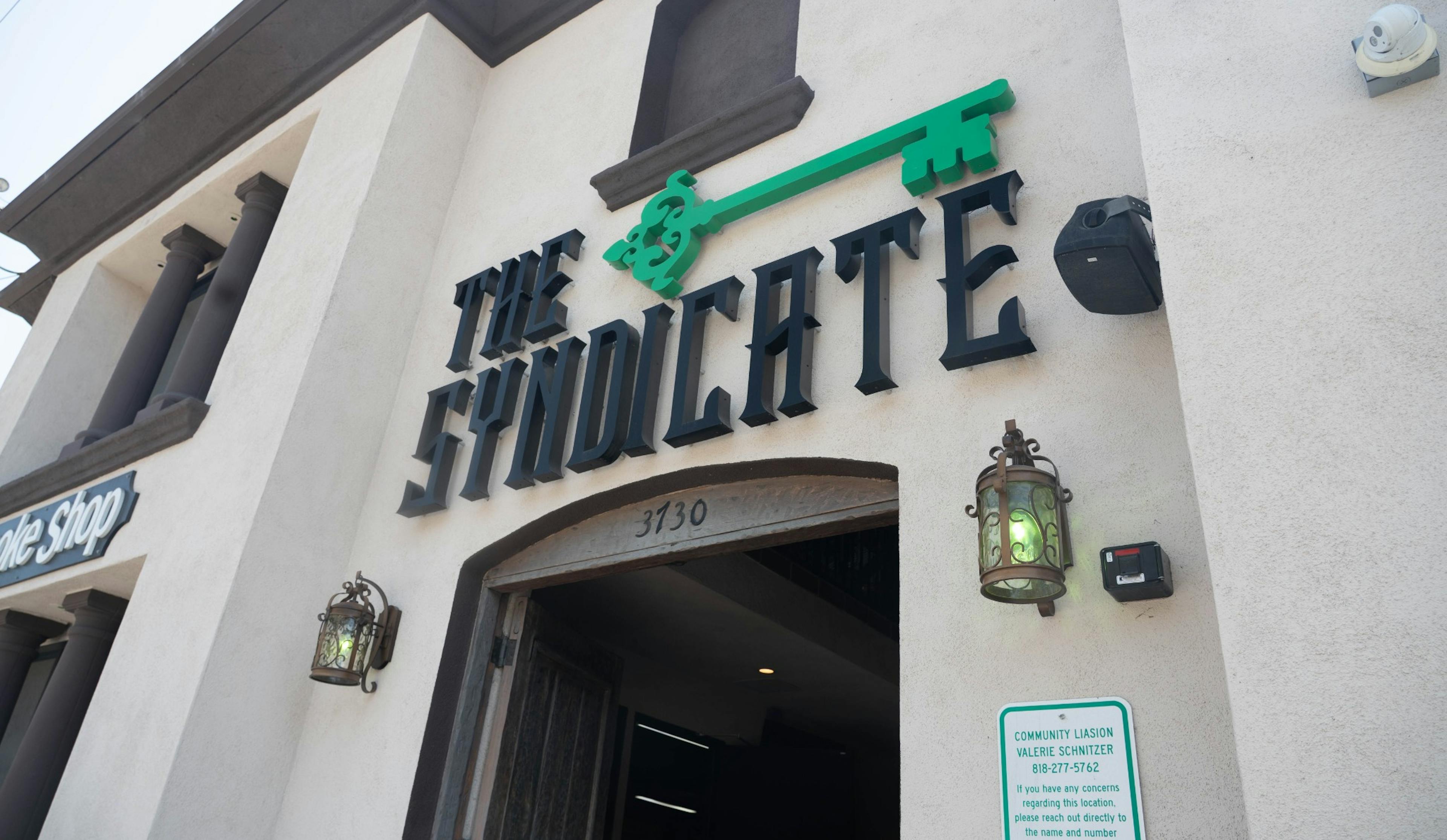 Angled upward view of The Syndicate Long Beach storefront showing the logo with green key and ornate lantern light fixtures above the door