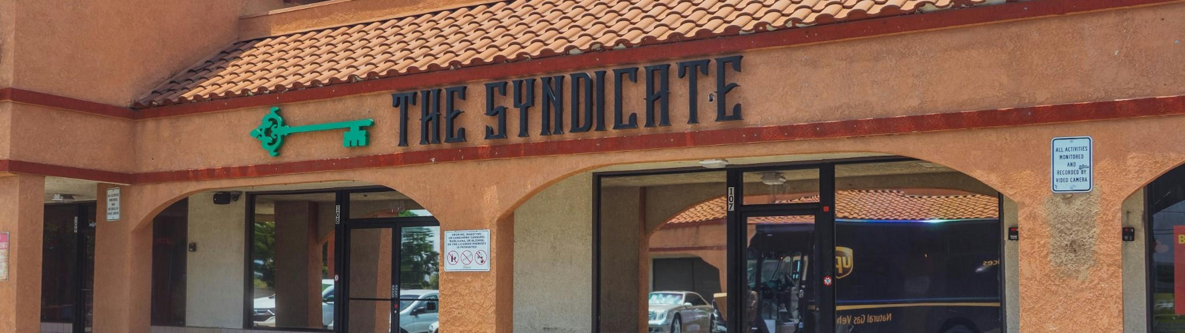 Street view of The Syndicate dispensary storefront with clear branding and signature green key logo in a SoCal plaza