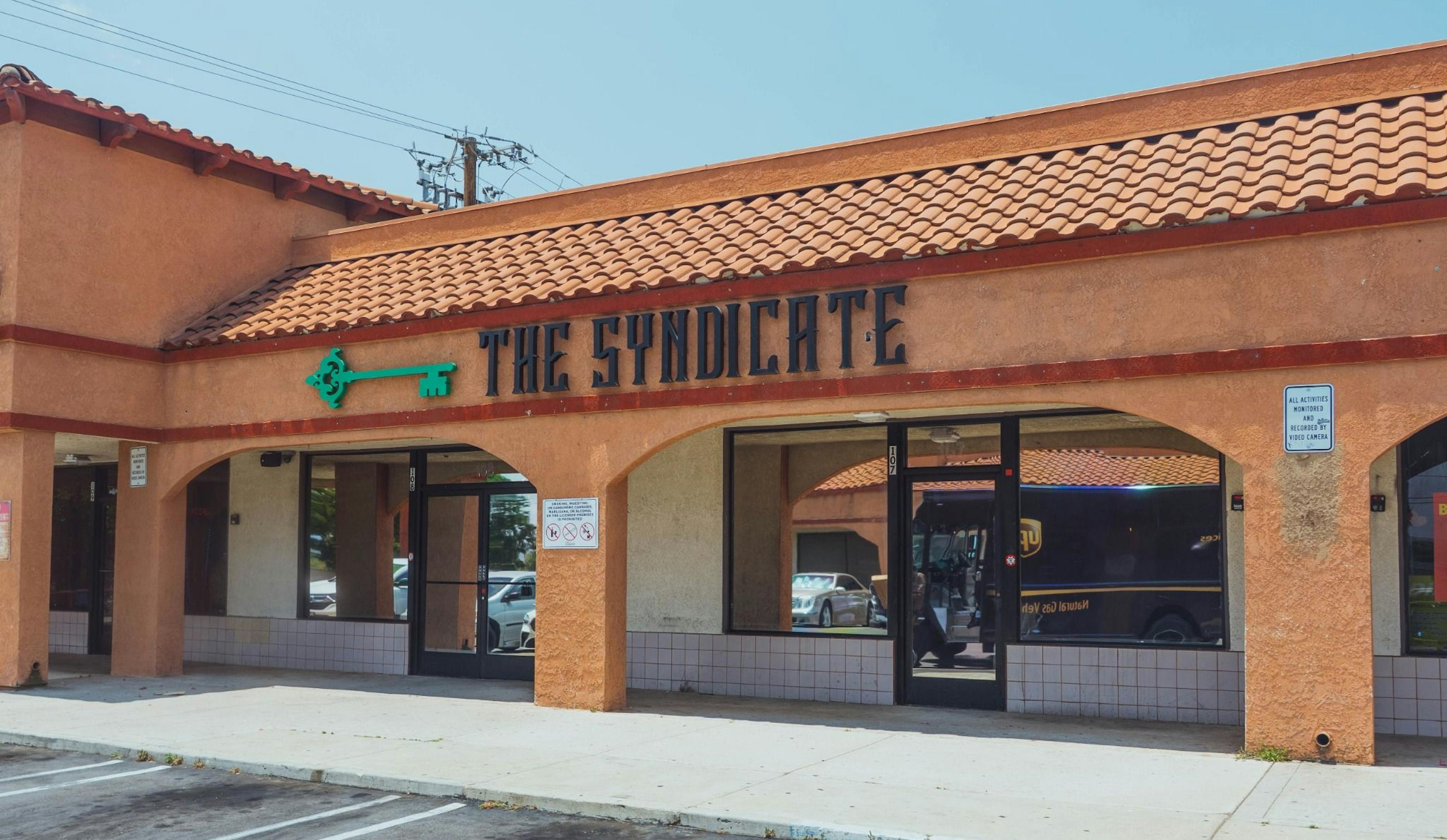 Street view of The Syndicate dispensary storefront with clear branding and signature green key logo in a SoCal plaza