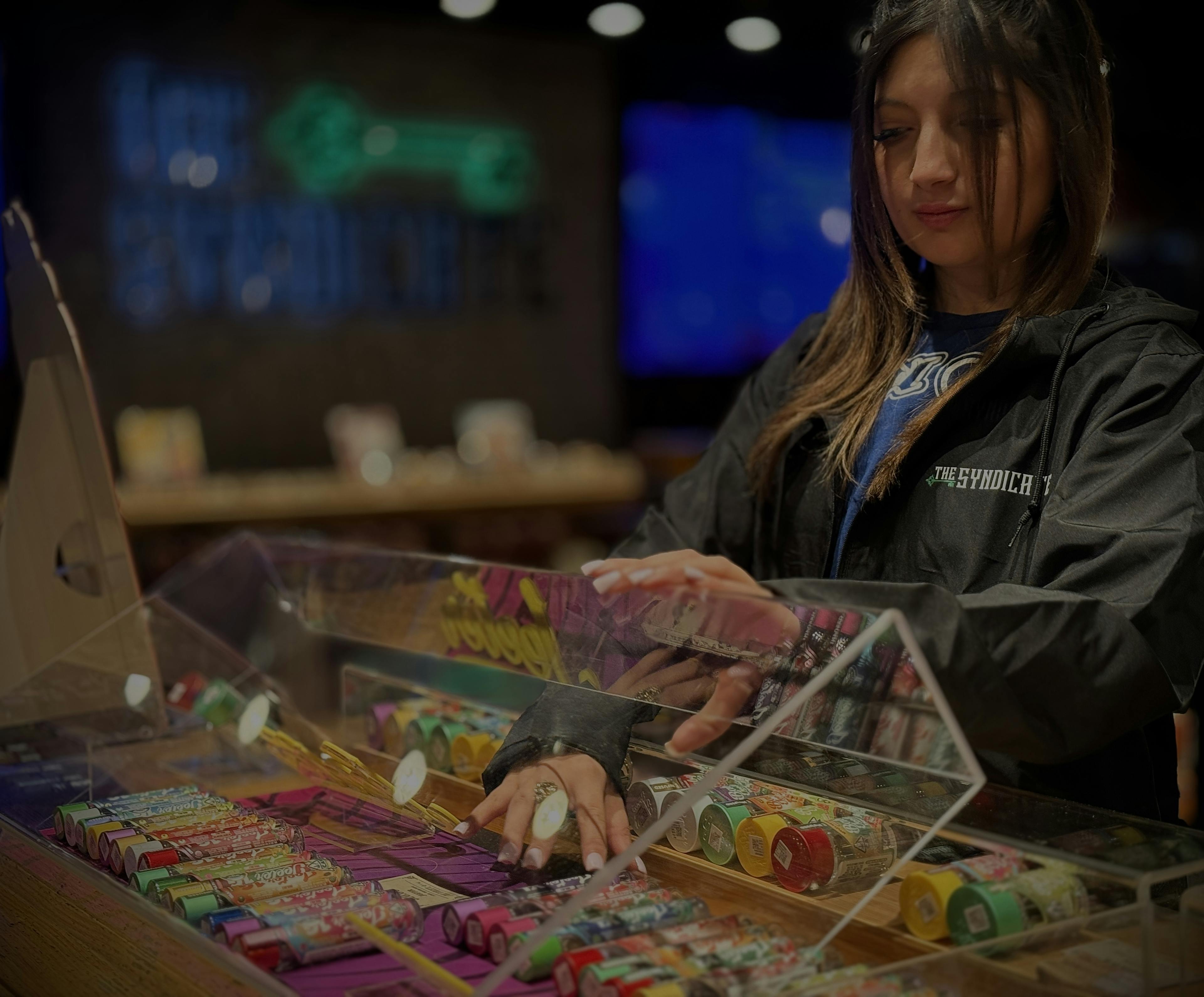 Female budtender at The Syndicate cannabis dispensary assisting customers and organizing pre-rolls and cannabis products in a modern retail display. Showcasing frontline retail career opportunities in California’s vertically integrated cannabis industry.