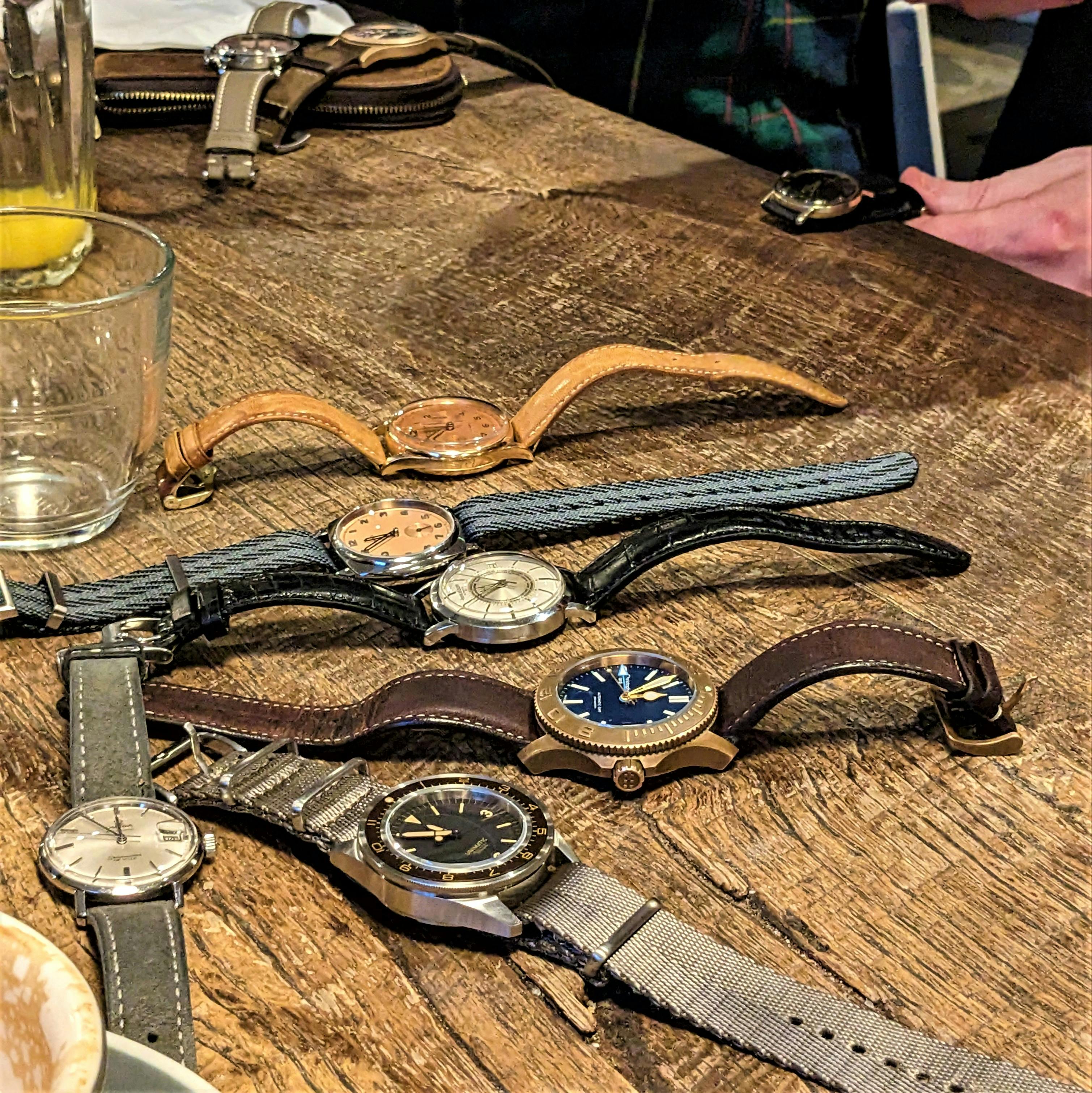 A pile of watches on a brown table featugin watches with differnet cases, coloured dials and straps. 
