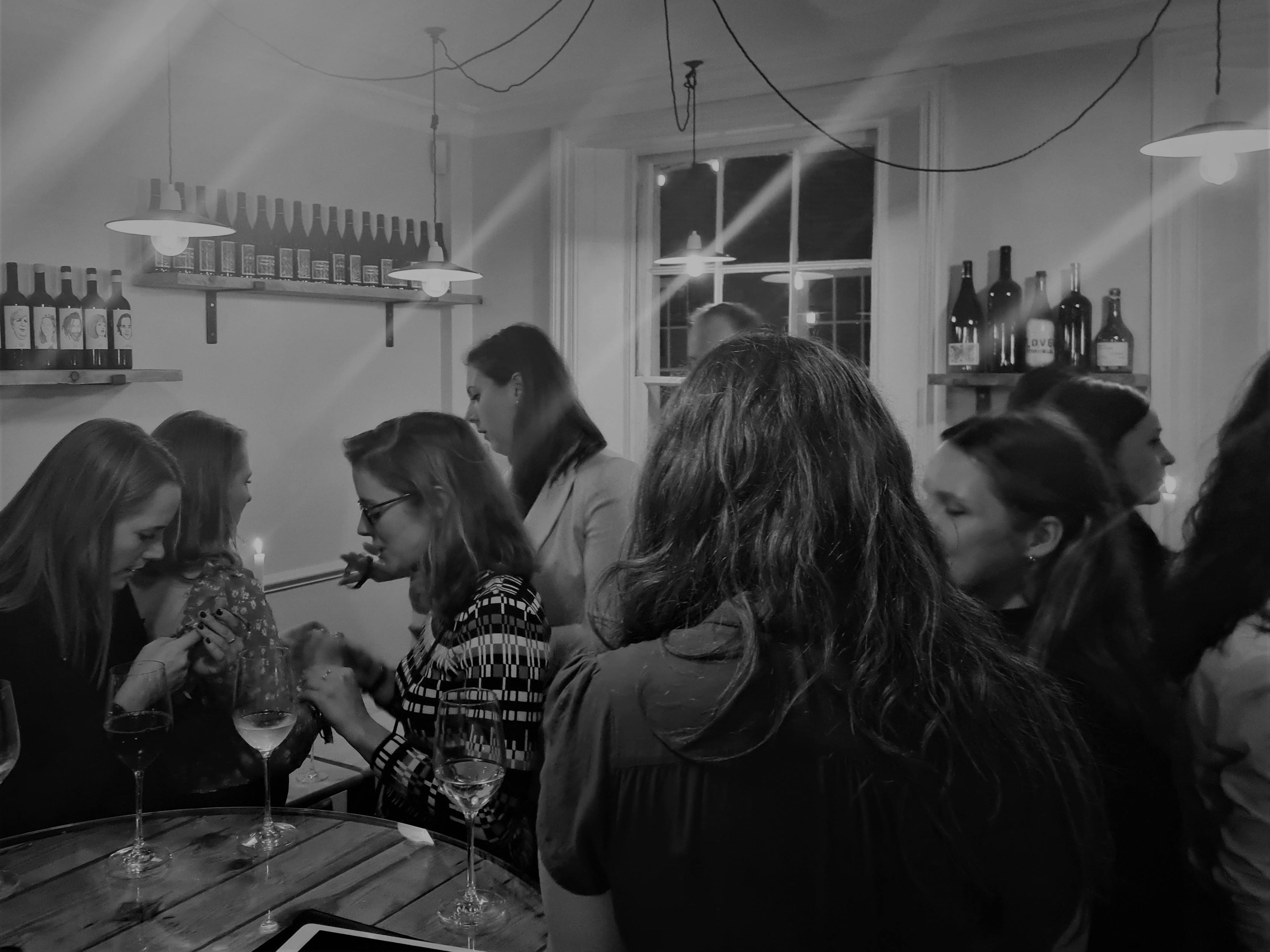 Black and white image of group of women talking in a wine bar with sparkling lights and wine bottles on shelves in the background