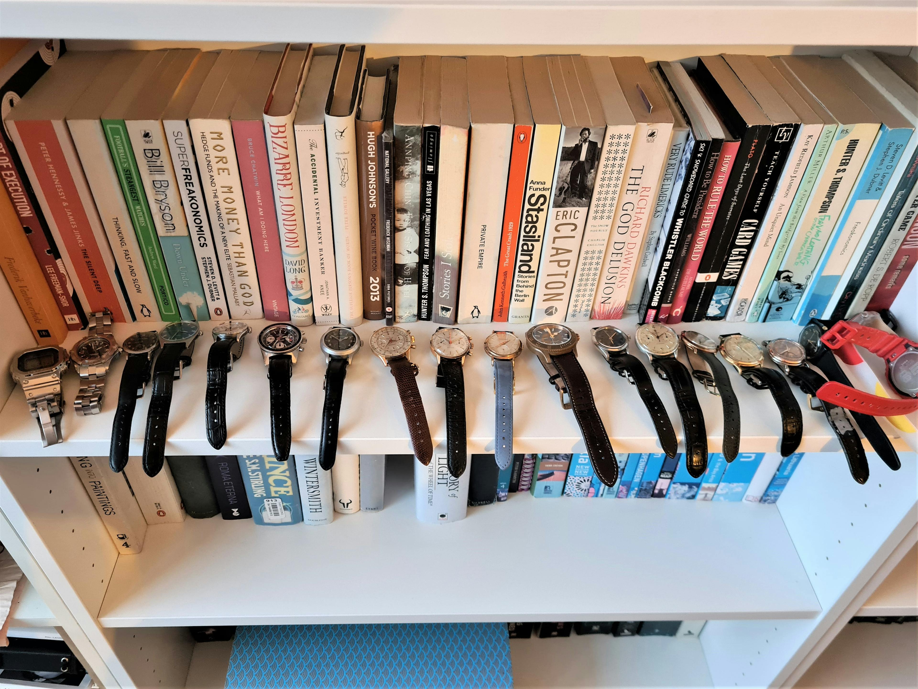 Over fifteen watches lined up on a bookshbelf, comprising a watch collection, with multiple types and straps.