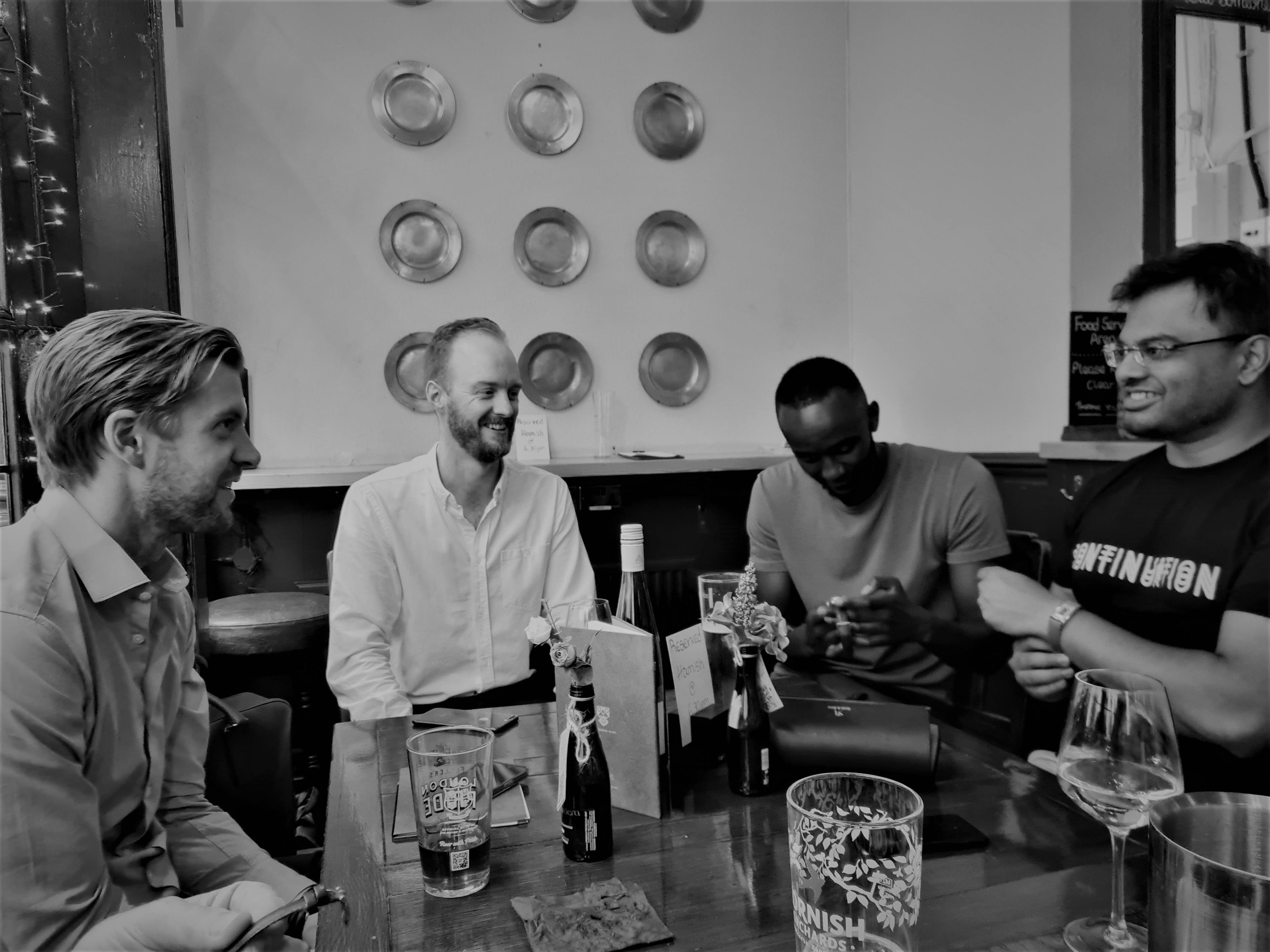 Four men laughing while looking at watches around a table in the pub, with drinks on the table and pewter plates on the white wall at the back.