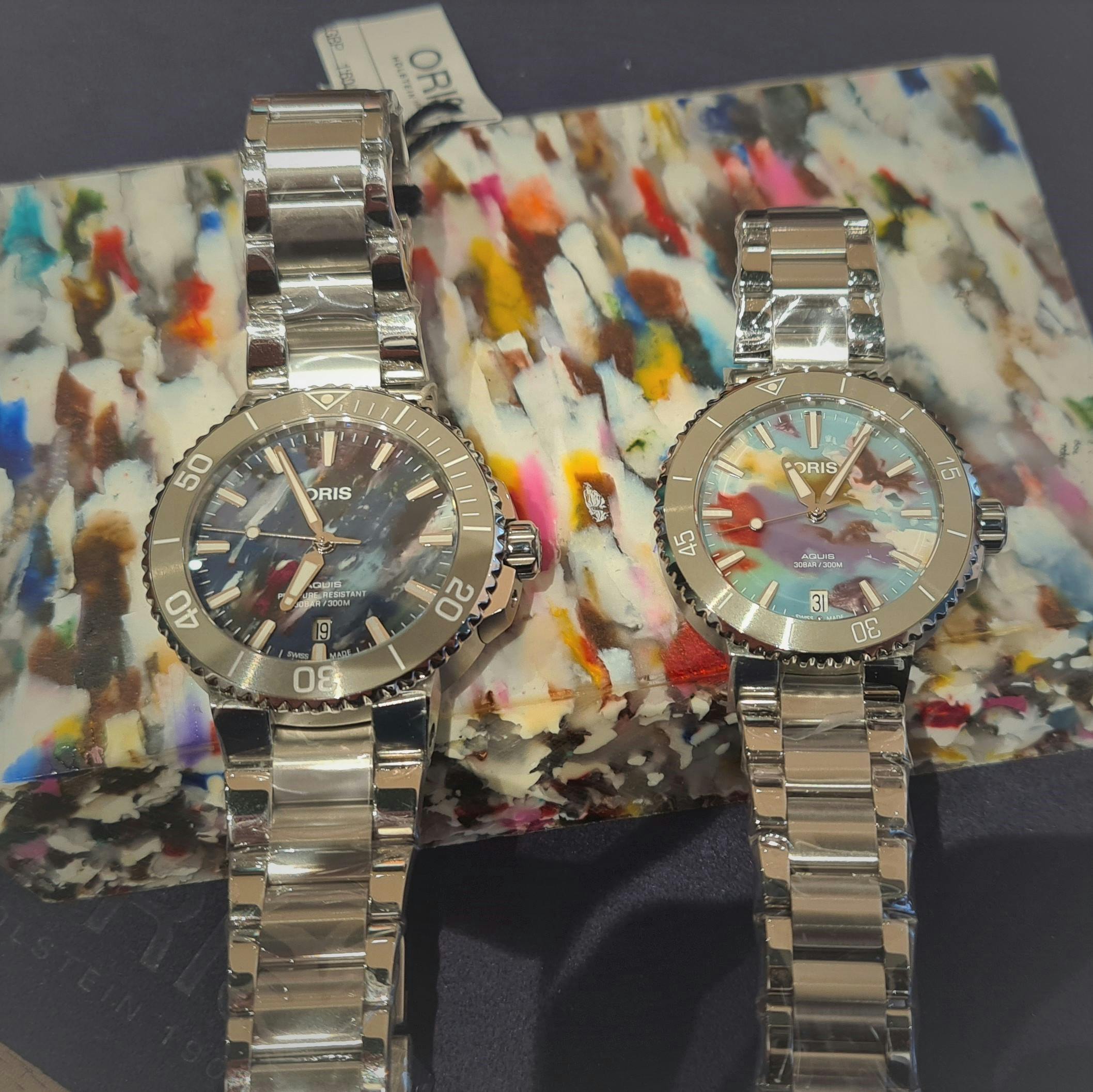 A Pair or Oris Aquis Upcycle watches on a block of mottle coloured plastic, both have steel cases and bracelets and inique multi coloured mottled plastic dials.