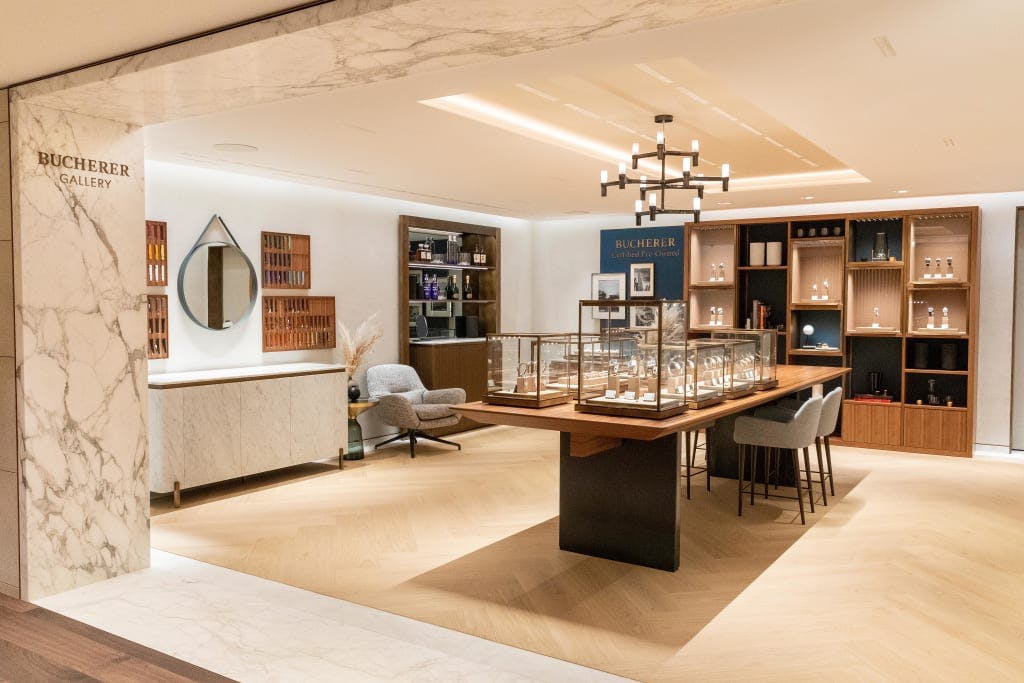 The interior of a luxury watch boutique with cream walls, wooden furniture including a shlving unit and table with watch display cases and a gold an white chandelier overhrad and a wooden floor at Bucherer Covent Garden