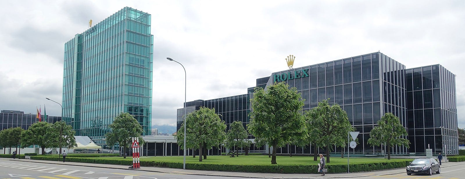 Rolex Factory in Geneva, featuring two office buildings one light green and one dark black with rolex logo on top and trees and grass in the foreground with grey sky above