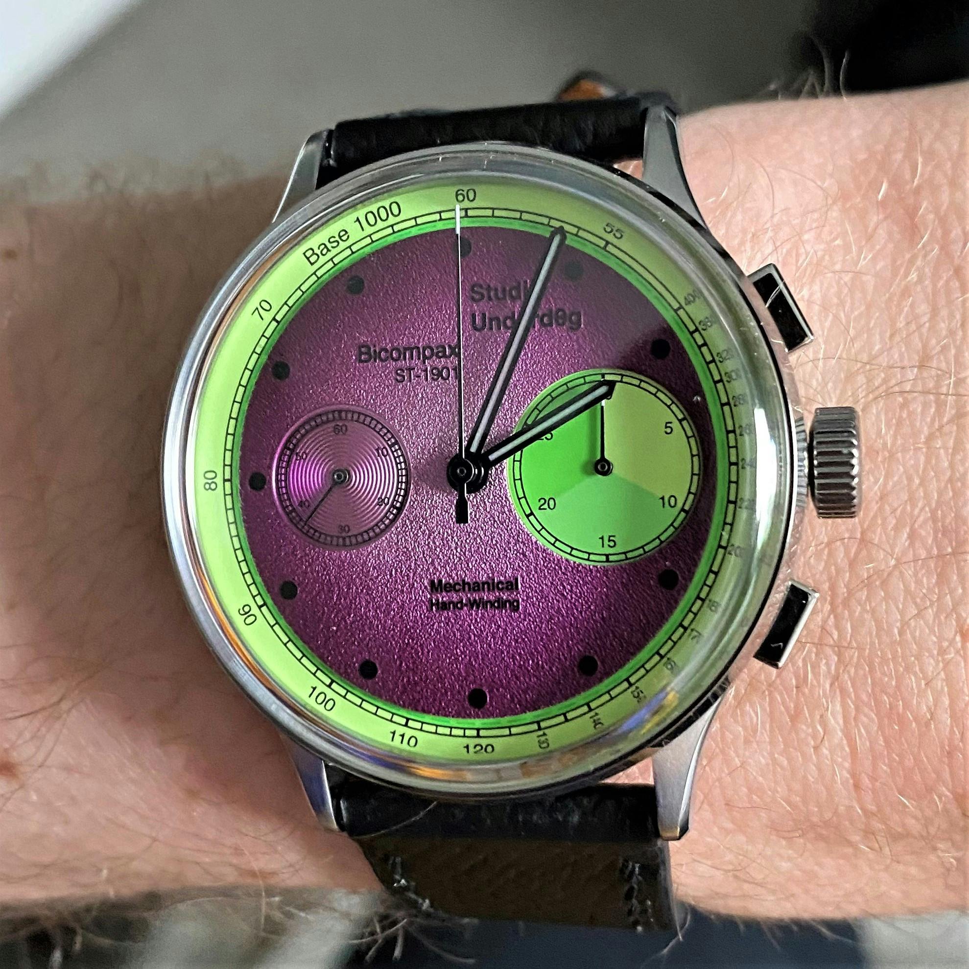 A Purple and Green Studo Underdog watch with two subdials, one large and one small, chronograph pushers on the right hand side and a luminous tahcnymeter track around the dial edge on a black strap. 
