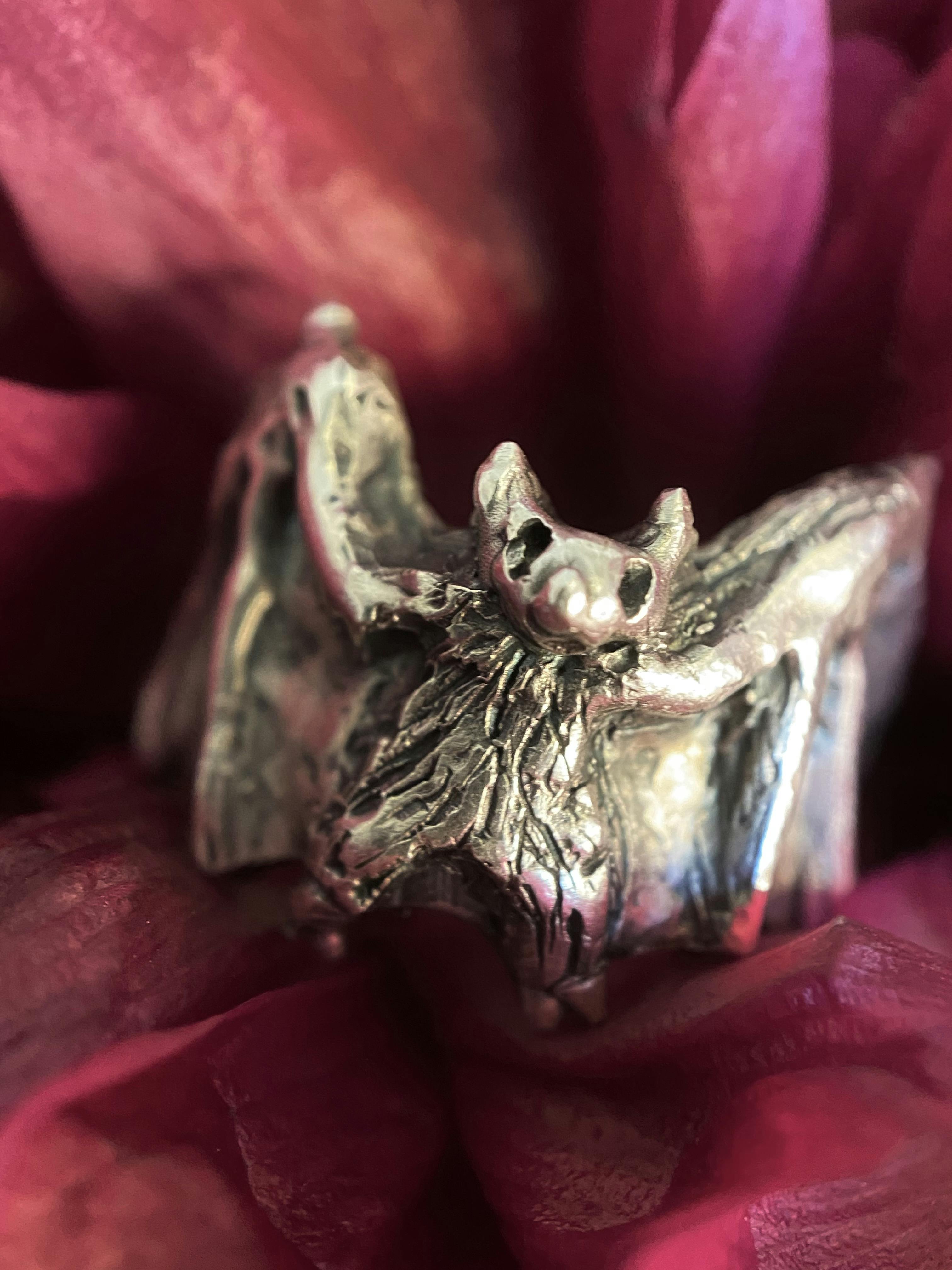 A small silver ring where the centre front shows he body of a bag, with black carved textural details and a little face. It’s wings wrap around the band to the back of the ring. 