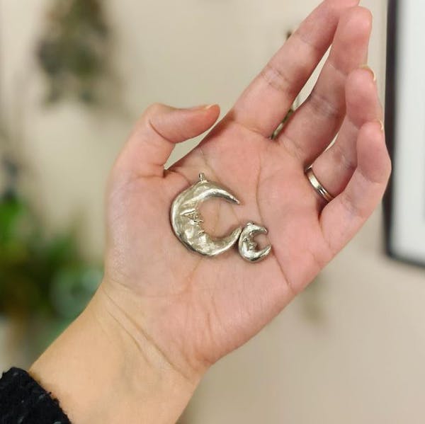 A large silver crescent moon face pendant and a much smaller one of the same design held up in the palm of a hand