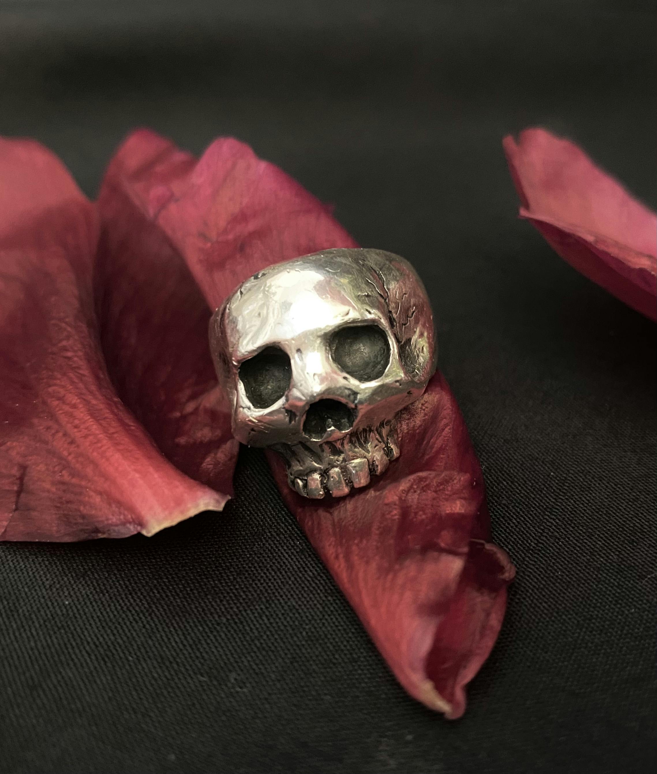 A large skull with no lower jaw made from silver sits on the front of the ring. The skull has blackened carved details of deep eye sockets, nose crevice and teeth, as well as some cracking details on the sides of the skull.