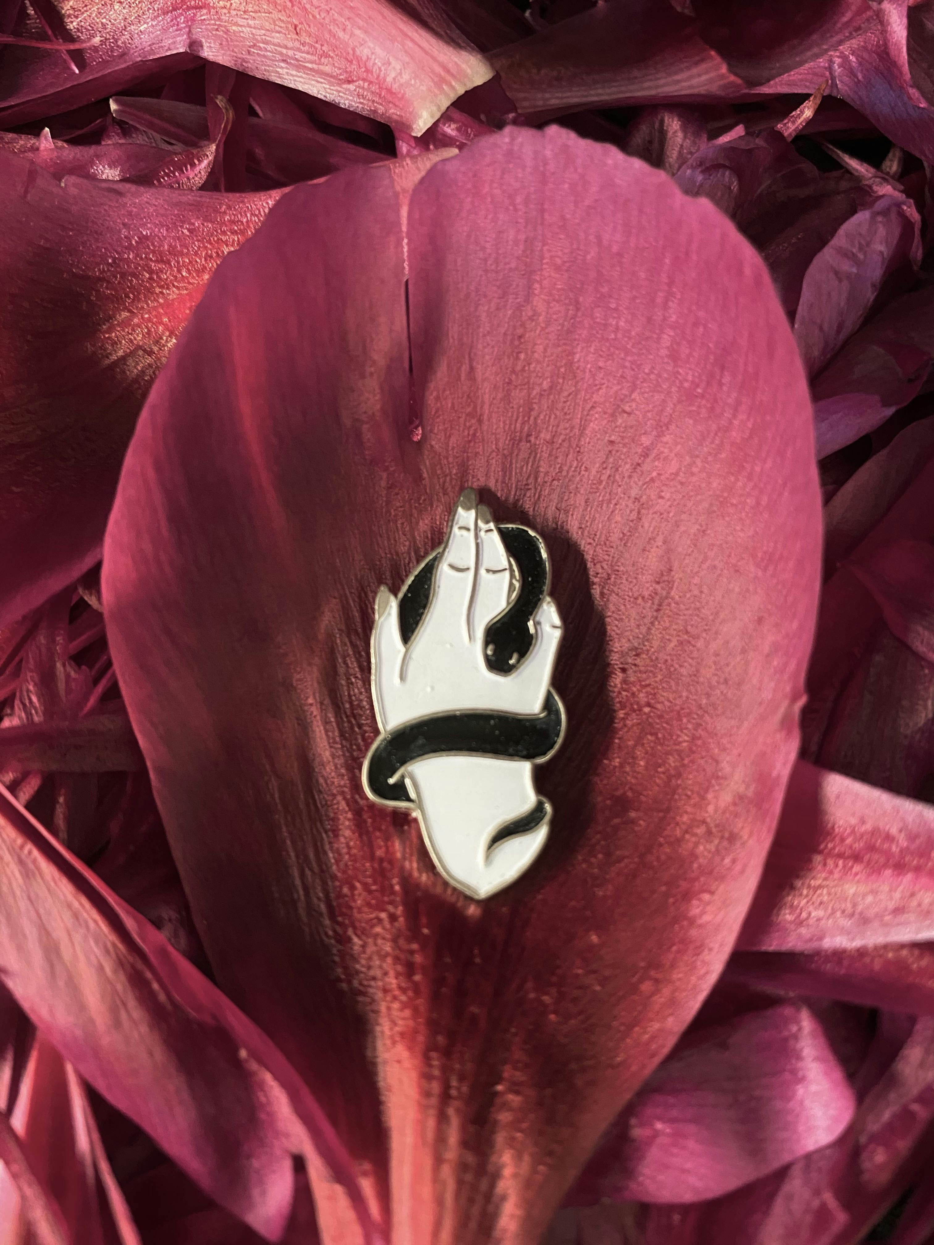An enamel pin in he image of a white hand with a black snake curled around it, where the lines around each element are made from silver metal. 
