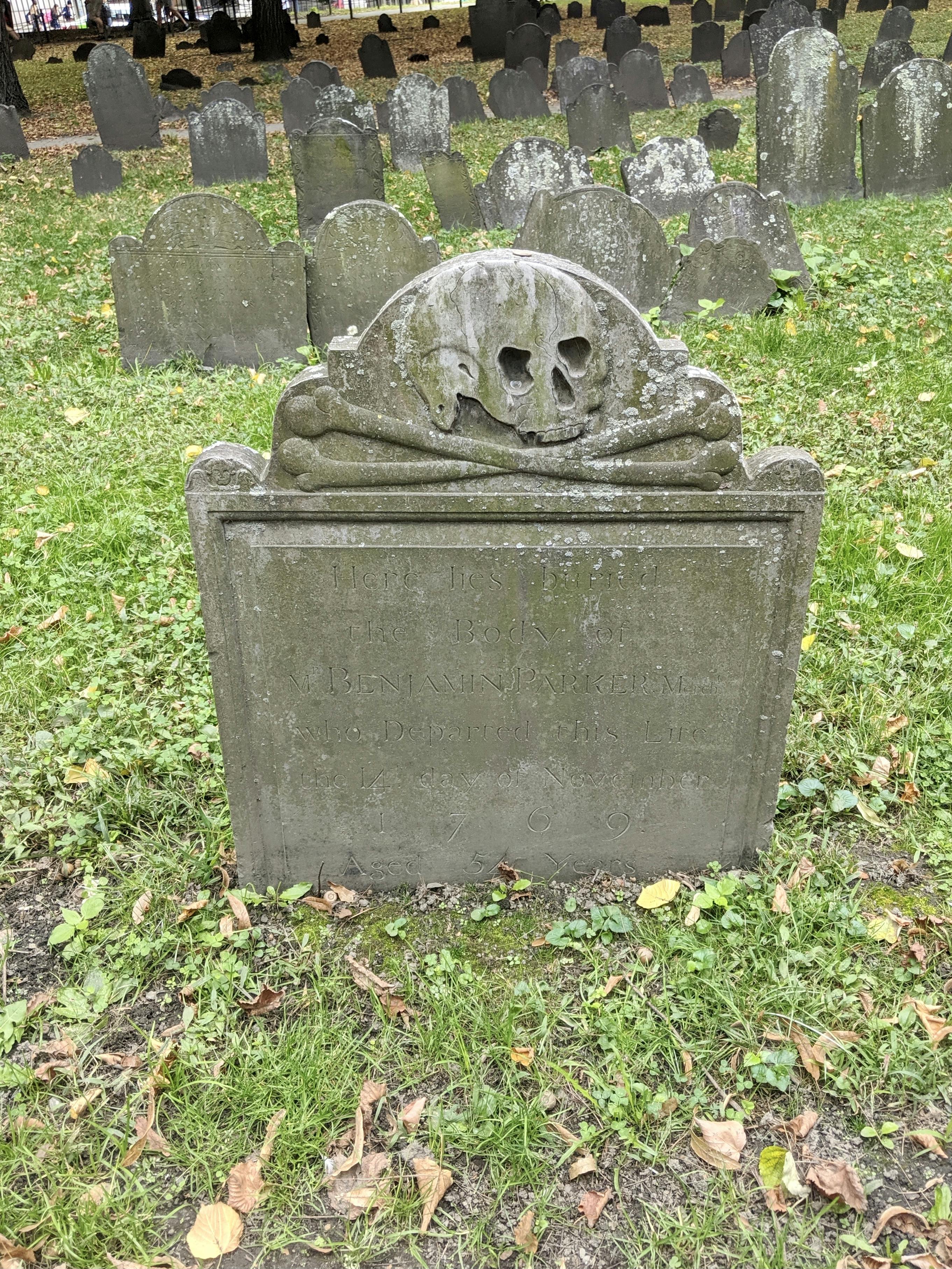 A photo in a graveyard, the stone in the foreground stands upright, at the top is a carved design showing a skull and crossbones. There is an inscription on the stone which is hard to read, but the date 1769 is clear.