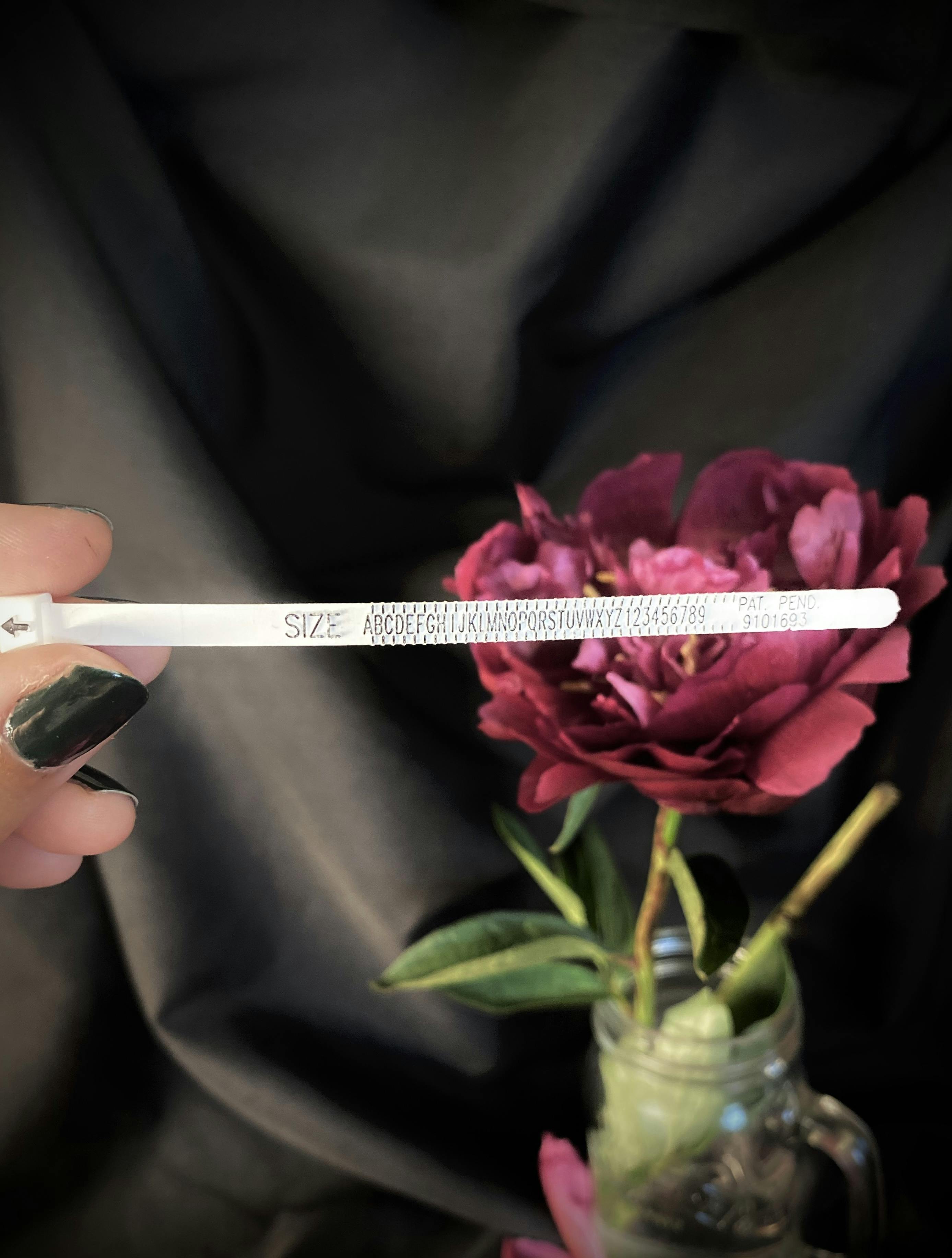 A hand holds up a thin strip of plastic with the word SIZE on followed by incremental measurement marks and the alphabet denoting ring sizes. In the background is a pink flower 