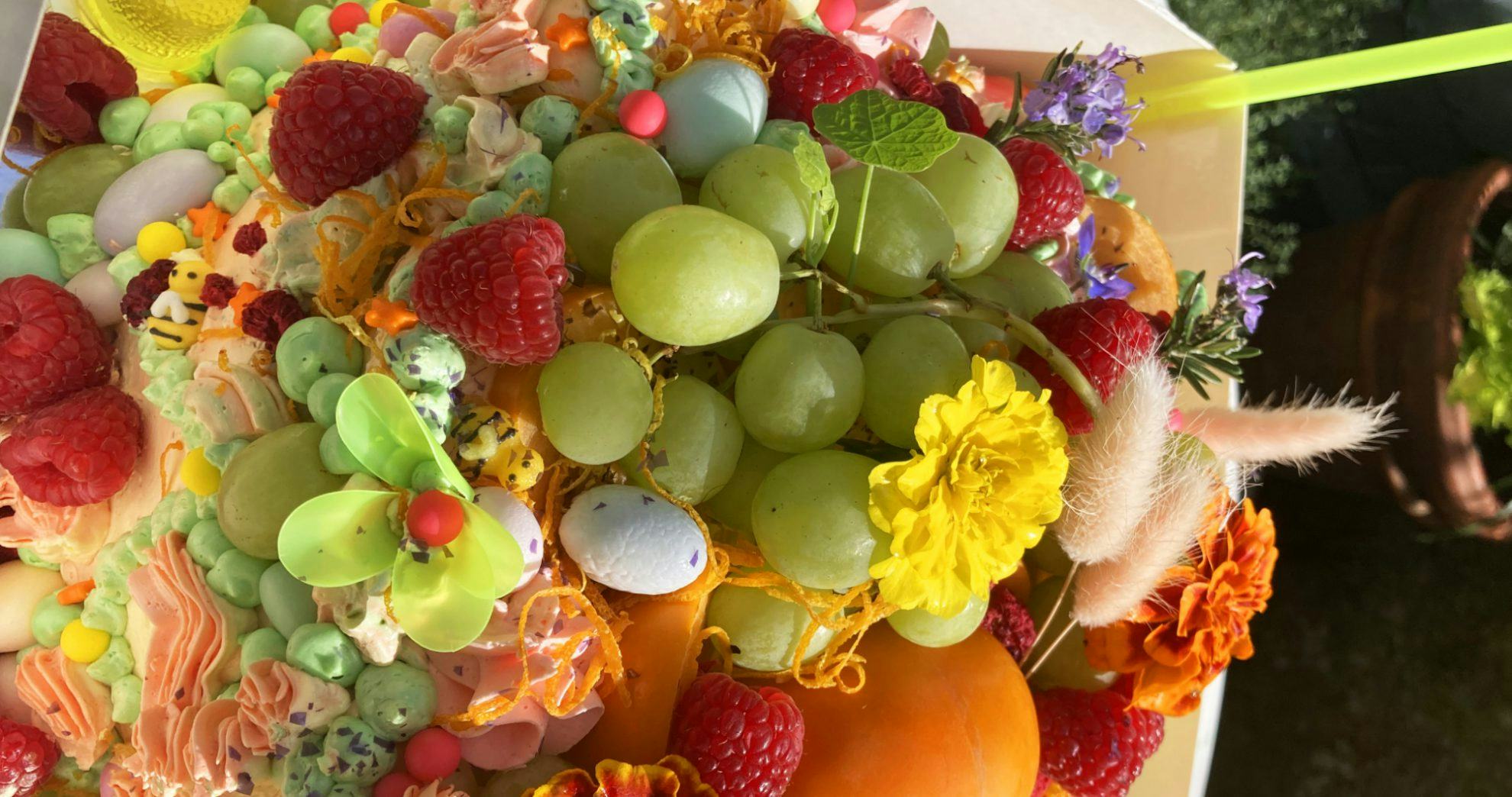 A cake close-up piled with peach and pale green icing, candy bumblebees and stars, grapes, raspberries, eggs, orange zest and flowers