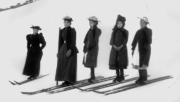A black and white photograph of young women skiing in full-length dresses and hats