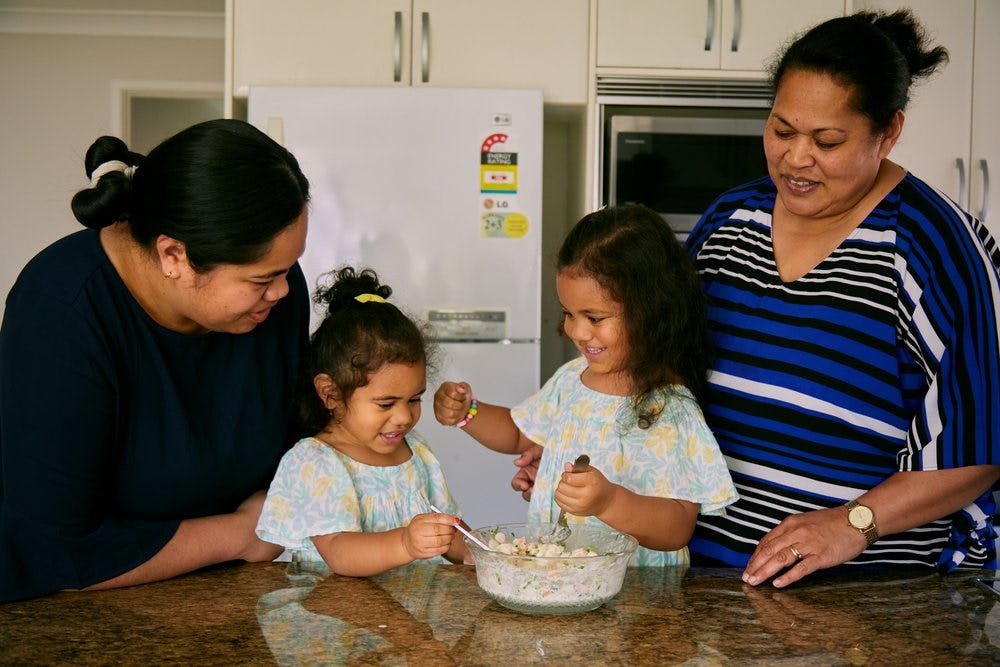 Sela Atiola stands to the left of her kitchen bench with her daughters in the centre and her mother to the far right. They make 'ota ika together with smiles on their faces. Photograph: Sherry Zheng. 