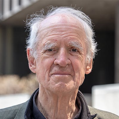 A portrait-style image of an older man with white hair. He is looking directly at the camera with a neutral expression. He wears a dark shirt and jacket, with blurred buildings and greenery in the background.
