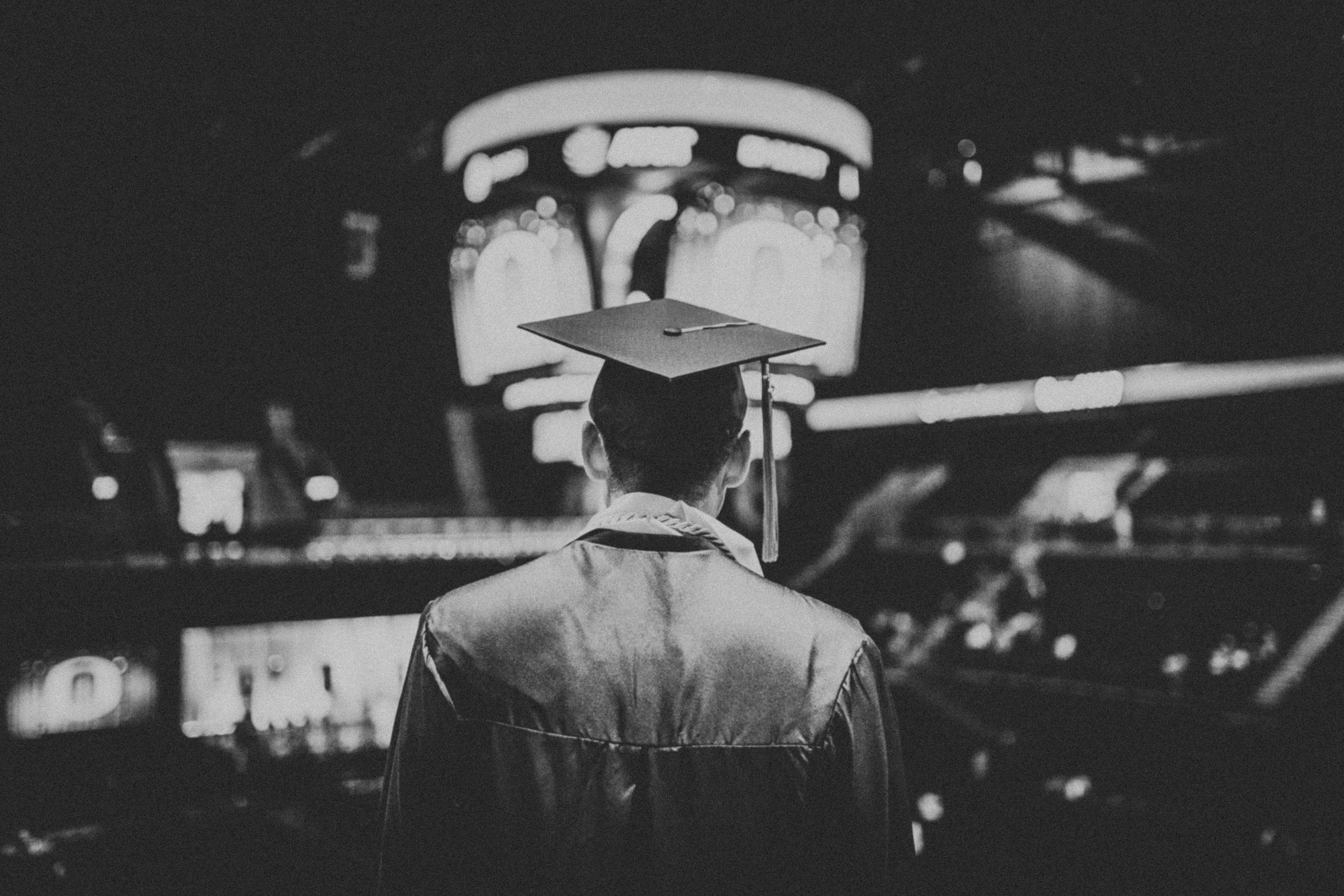 A black-and-white image of a graduate standing with their back to the camera, wearing a graduation cap and gown, looking out into a large, dimly lit auditorium with glowing displays in the background, capturing the moment of reflection during a graduation ceremony.