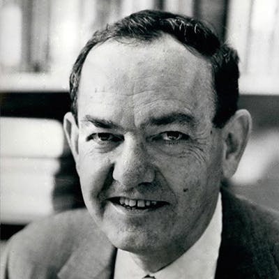 A man is smiling while wearing a suit and tie in a portrait photograph; the background shows a blurry library filled with books.
