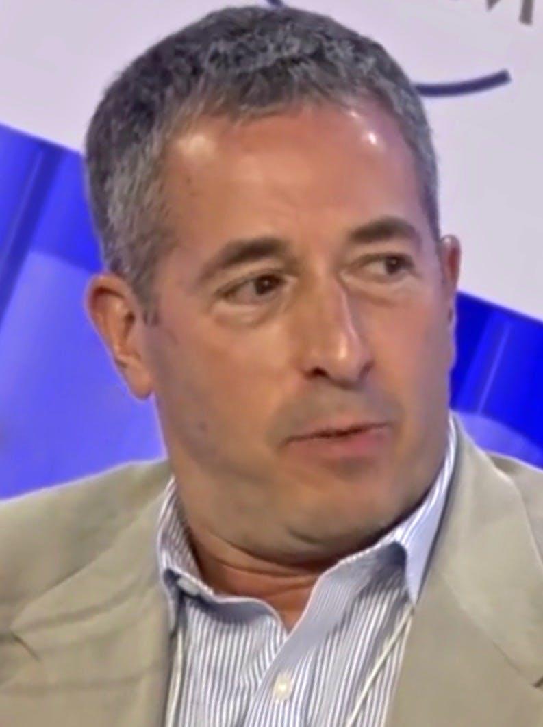 A man with short gray hair and a light-colored suit is speaking on a panel, with blue and white backdrop elements visible behind him.