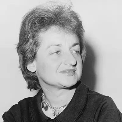 A person with short, tousled hair and a thoughtful expression looking to the side, wearing a dark top and a necklace with a pendant, against a plain background.