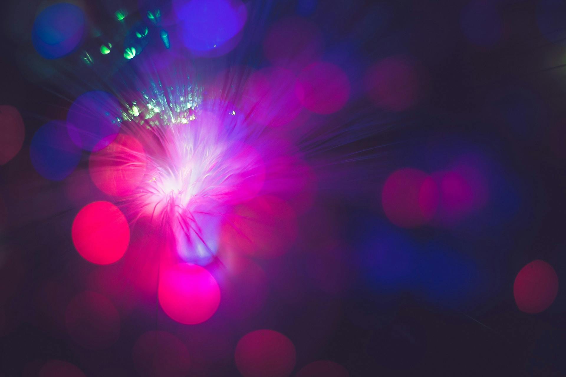 An abstract burst of light with pink, purple, and blue hues radiating outward, resembling a blend of soft bokeh effects and energetic light streaks against a dark background.