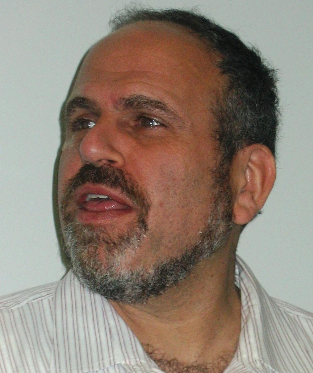 A bearded man with short hair is looking to his left with an open mouth, as if speaking, against a plain light-colored background. He wears a striped shirt.