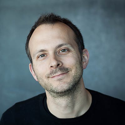 A man is smiling slightly, facing the camera, wearing a black shirt, with a grey, blurred background.