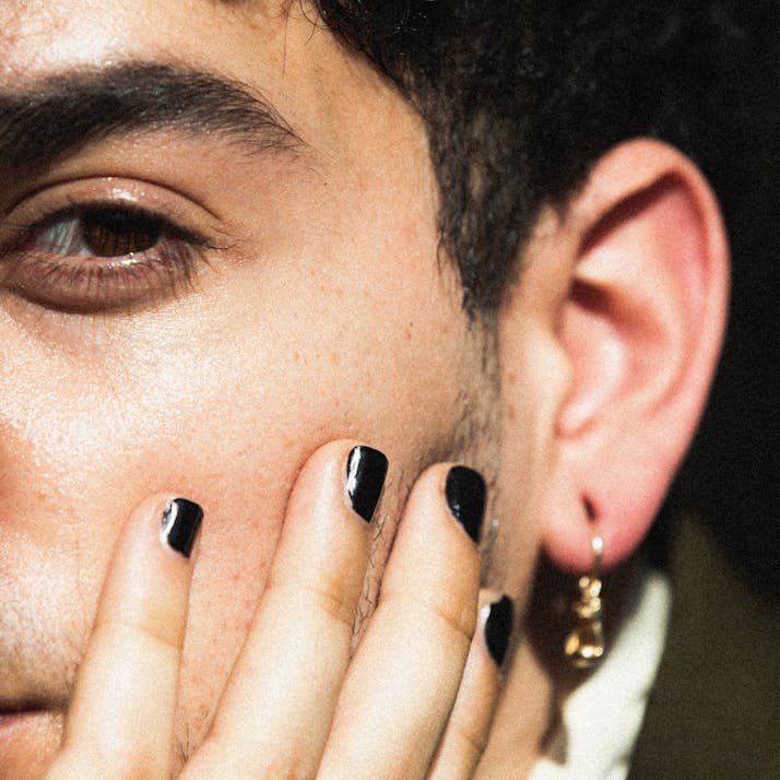 A man with his hand held up to his face, nails painted black, staring into camera.