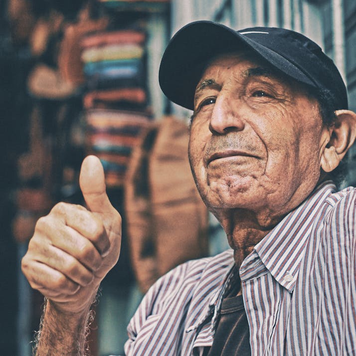 An older man wearing a baseball cap gives a thumbs up.
