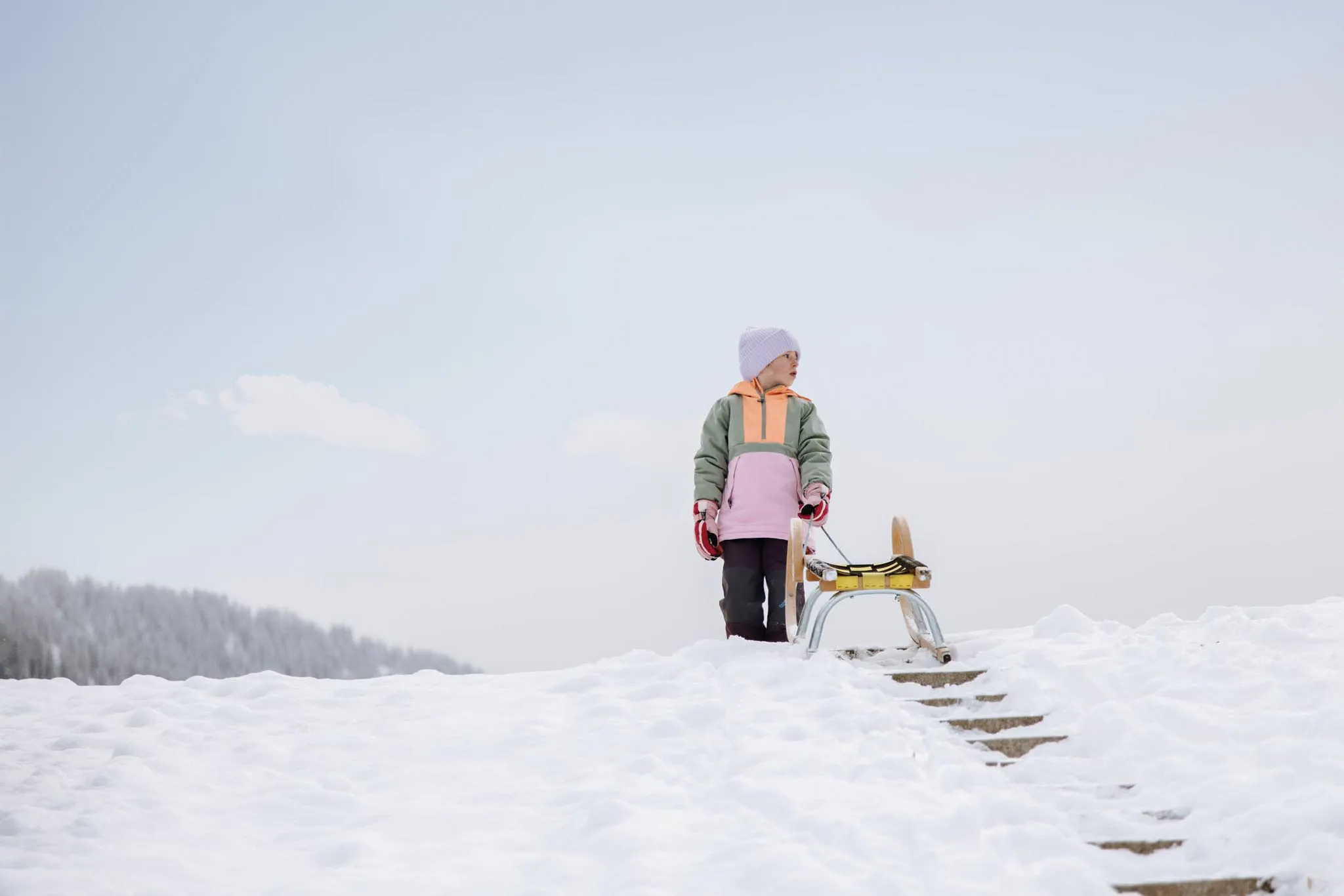Girl standing with a sledge in the snow in the hotel gardens of the Dachsteinkönig – Familux Resort.