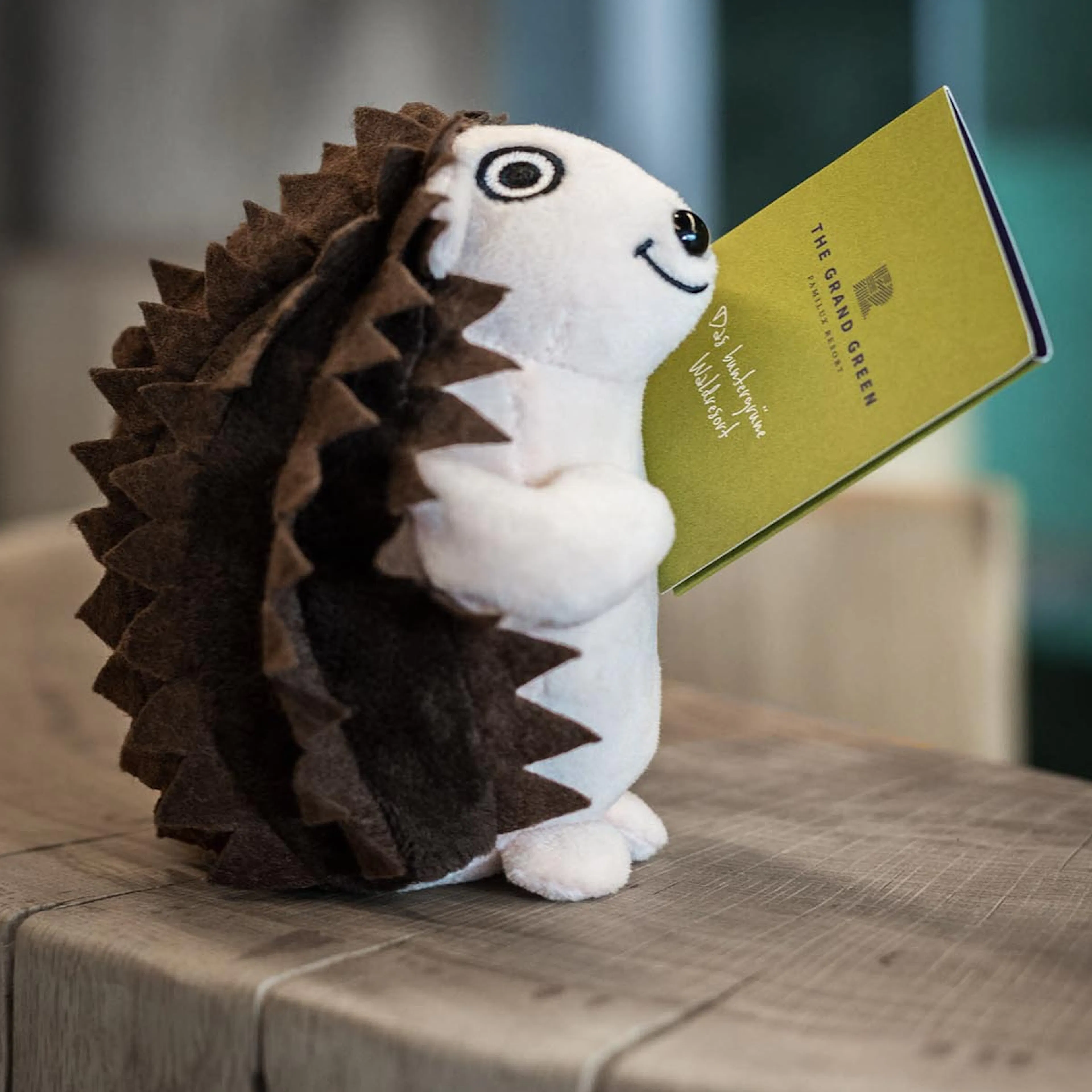 Maskottchen Igel Iknazz sitzt auf dem Rezeptionstresen mit einer Zimmerkarte des The Grand Green - Familux Resorts.