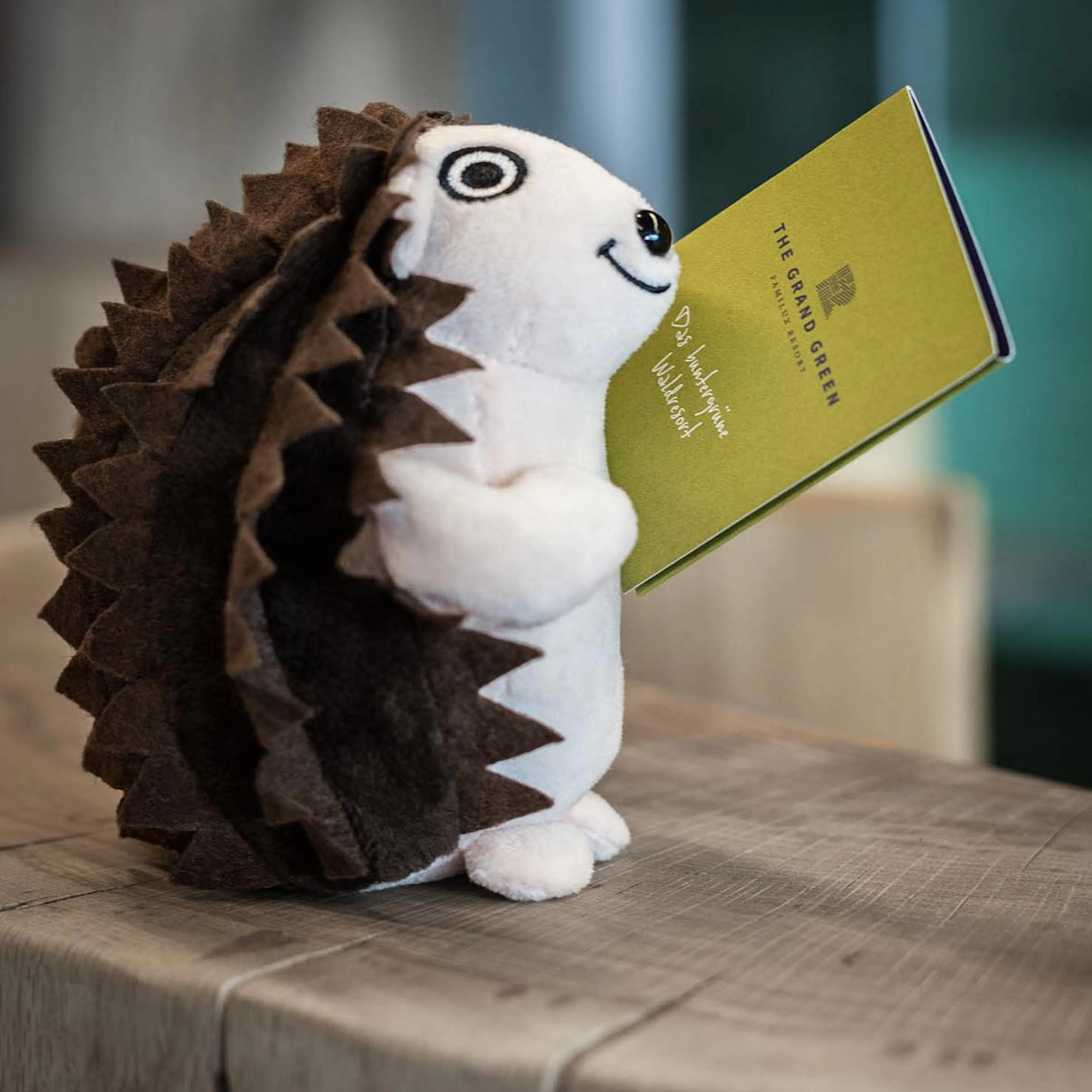 Maskottchen Igel Iknazz sitzt auf dem Rezeptionstresen mit einer Zimmerkarte des The Grand Green - Familux Resorts.