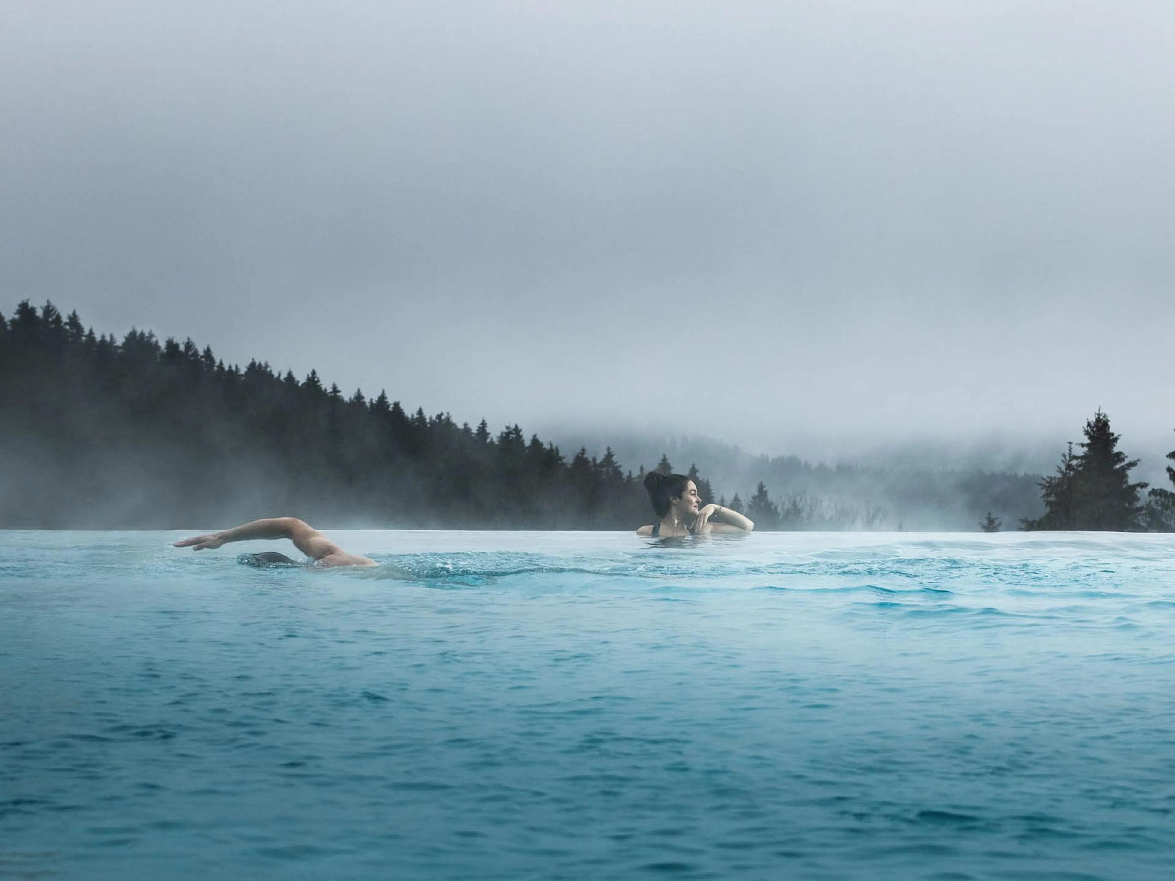 Woman and man swimming in the outdoor pool at The Grand Green – Familux Resort with views into the forest.