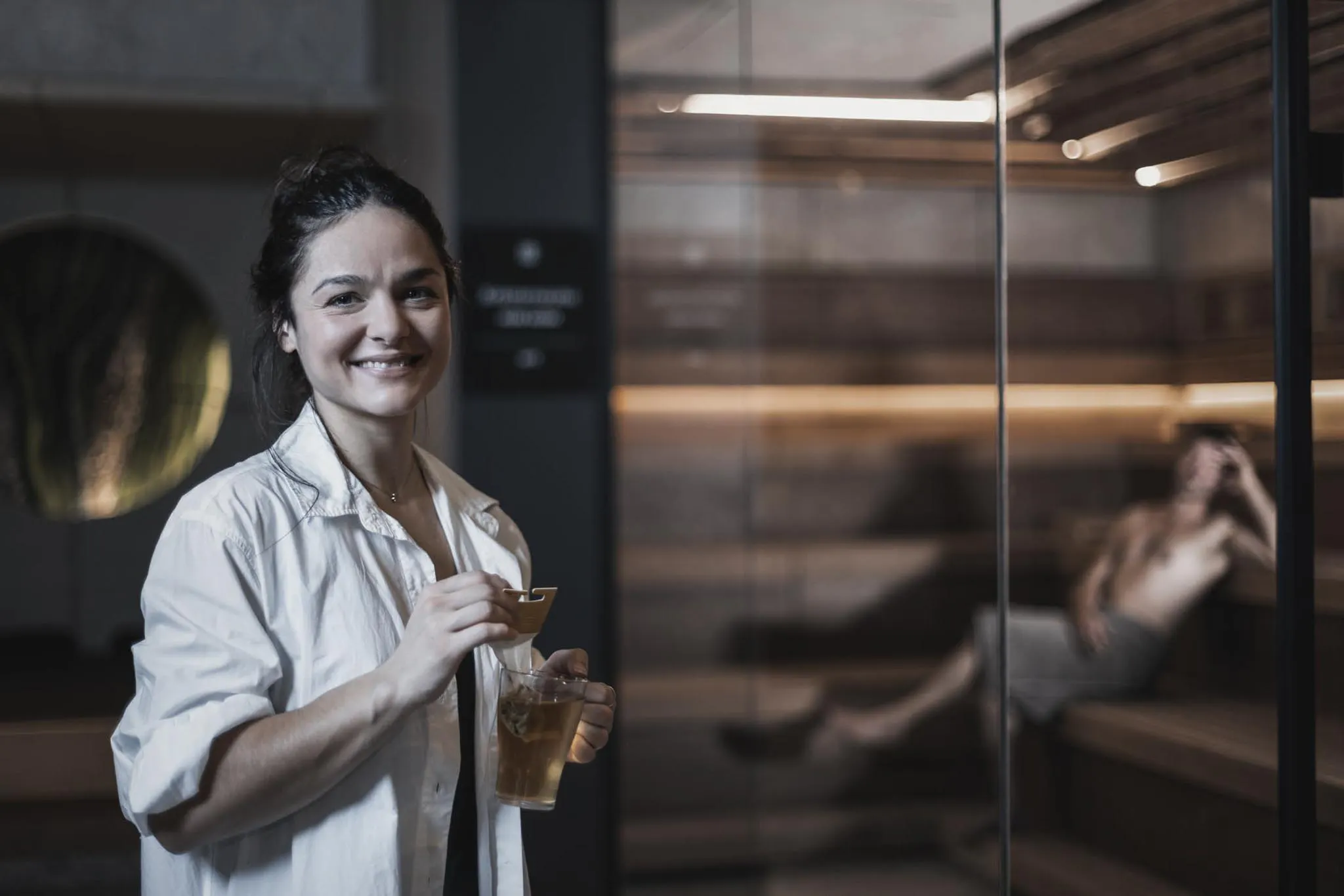 Frau steht mit Tee vor der Sauna im Wellnessbereich des The Grand Green - Familux Resorts.