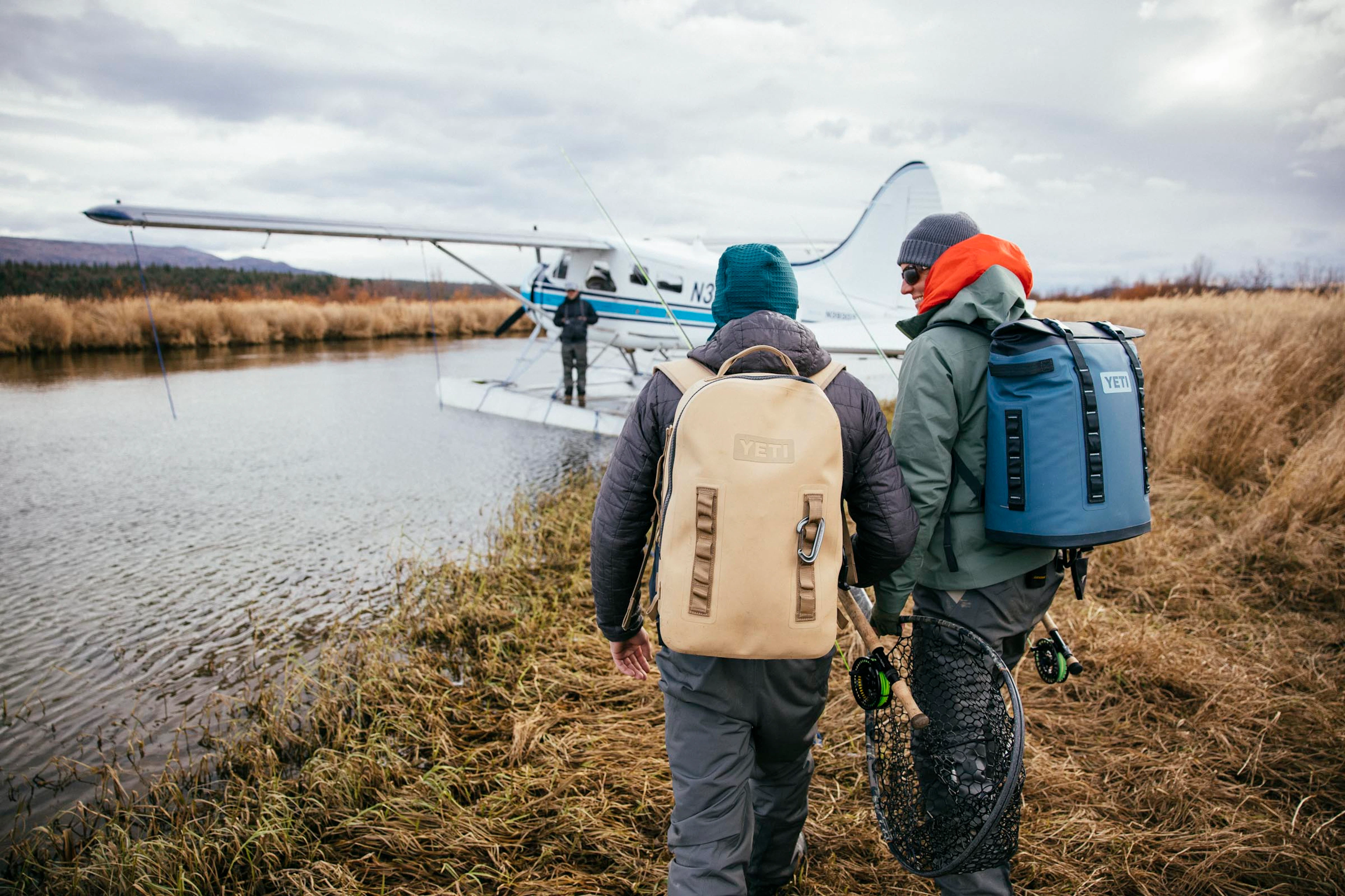 Anglers carrying fly rods and gear along the riverbank toward a float plane for a fall fly-out fishing trip in Katmai National Park, Bristol Bay, Alaska
