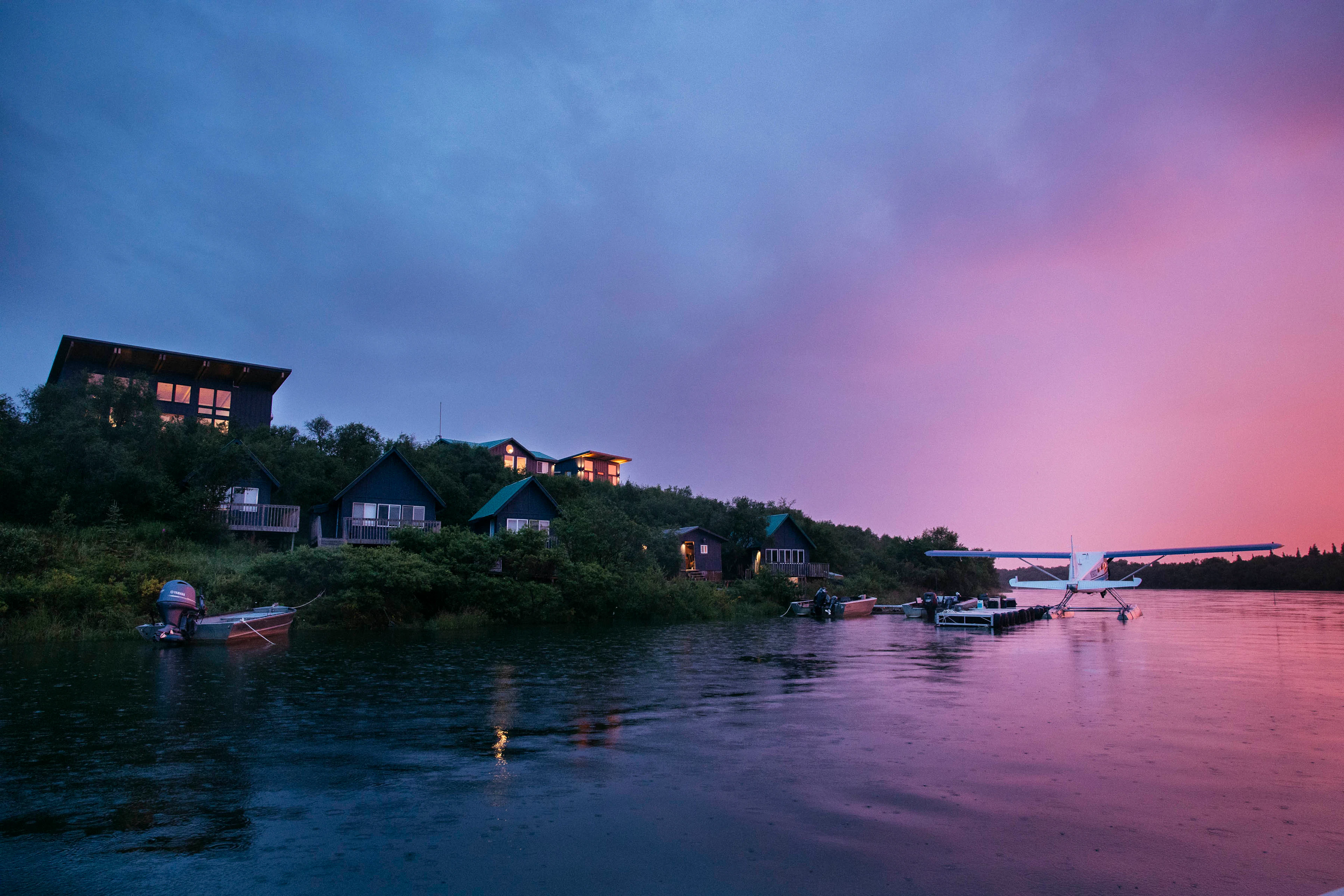 The Lodge at 58* North at dusk, seen from the Naknek River, with the main lodge and guest cabins lit up on the wooded bank, fishing boats tied along the shore, and a floatplane at the dock under a pink and purple Alaska sky
