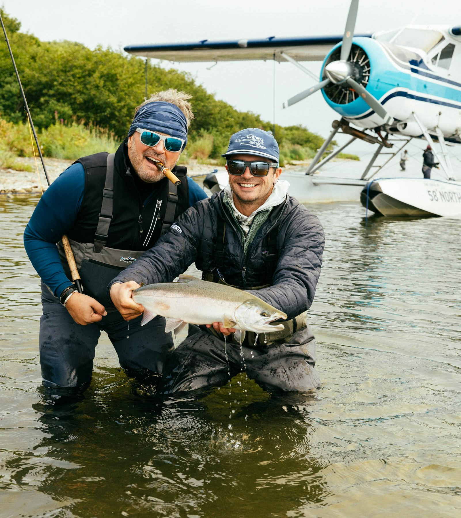 Two anglers holding a trophy fish in shallow water with an Alaska floatplane behind them on a guided fly-fishing trip