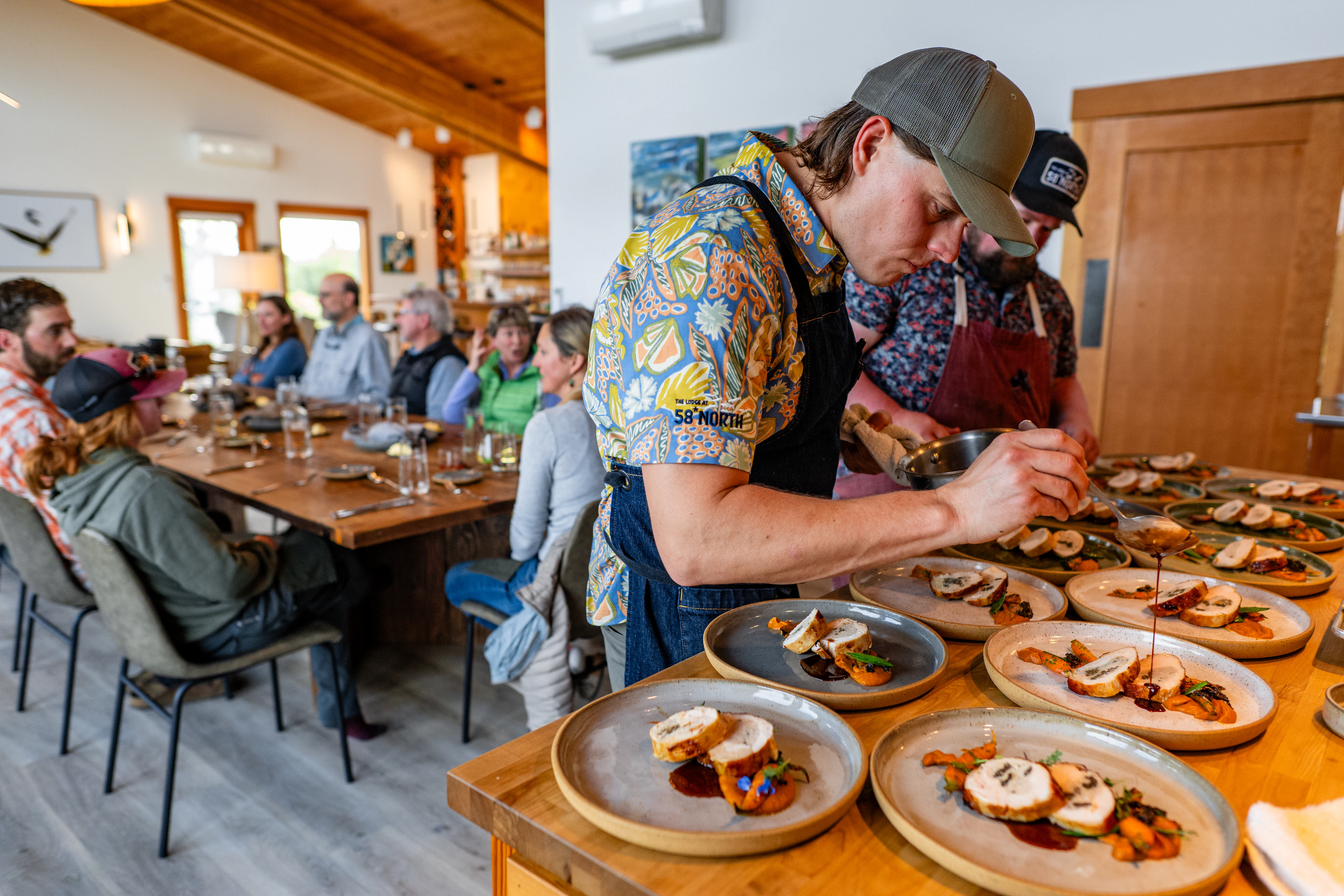 A chef plates dinner in the main lodge at The Lodge at 58* North while guests gather around the dining table in Bristol Bay, Alaska.