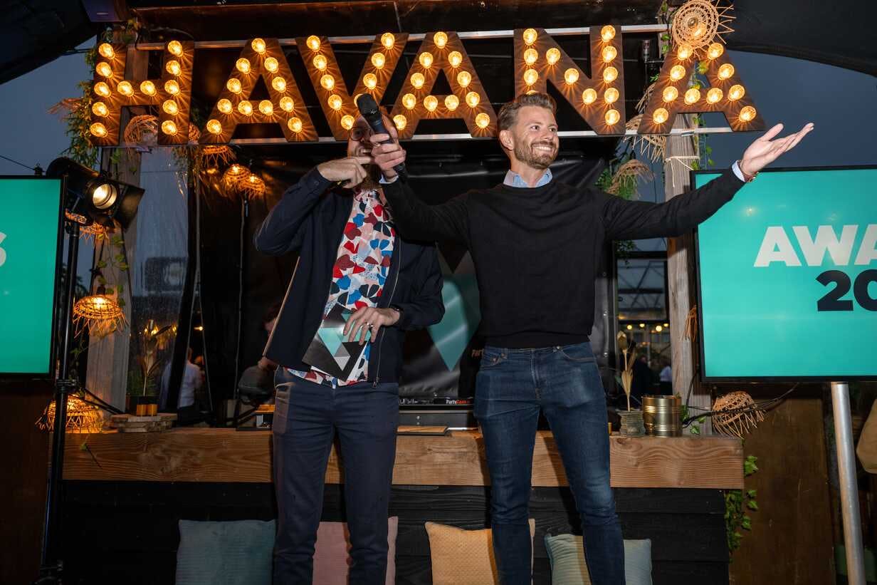 Raico van Elsen staat lachend me open armen en microfoon in de hand awards uit te reiken op het zomer event van The.NextGen samen met een presentator die half achter hem staat.